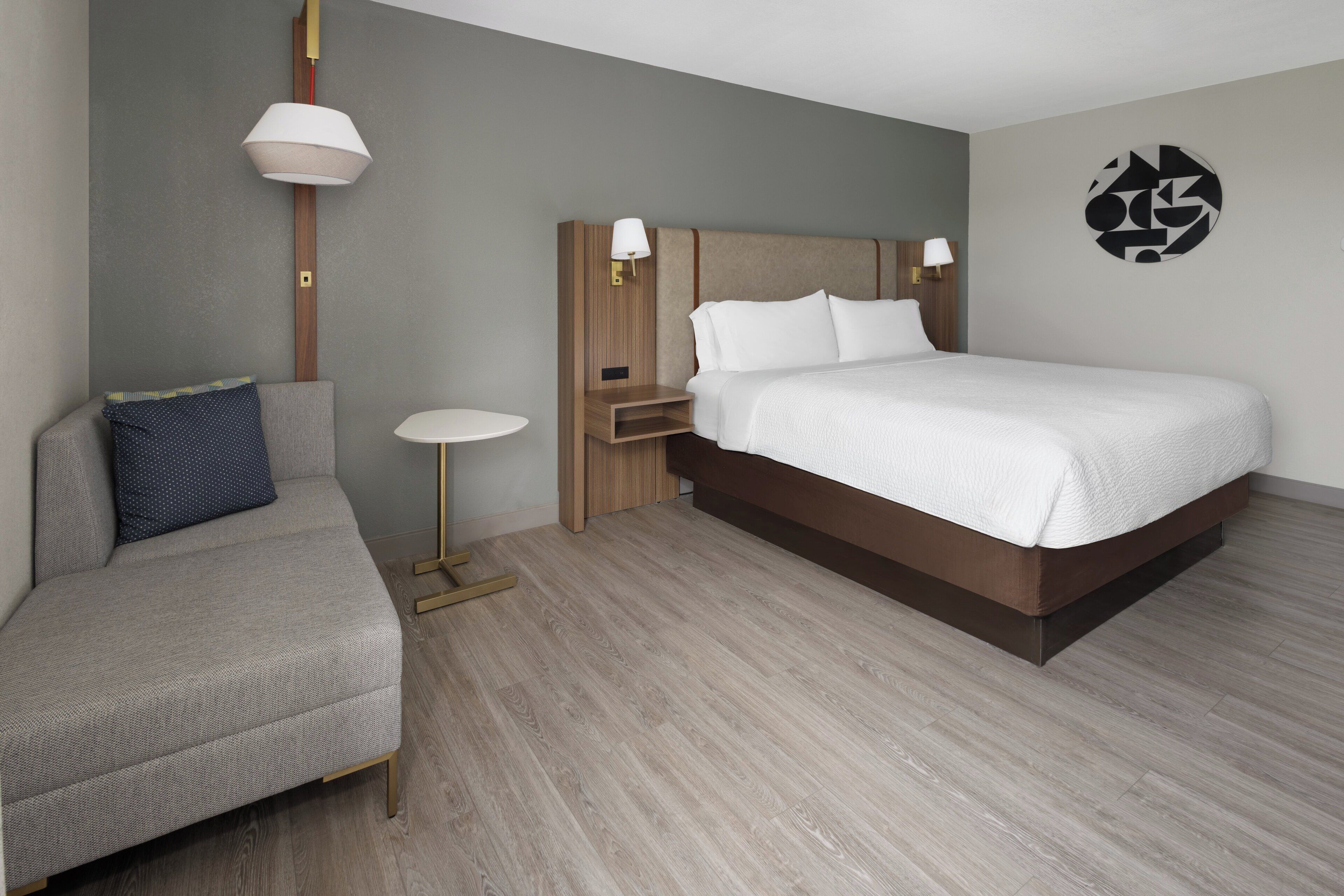A modern hotel room with a bed, a sofa, a small table, and a wall art piece.