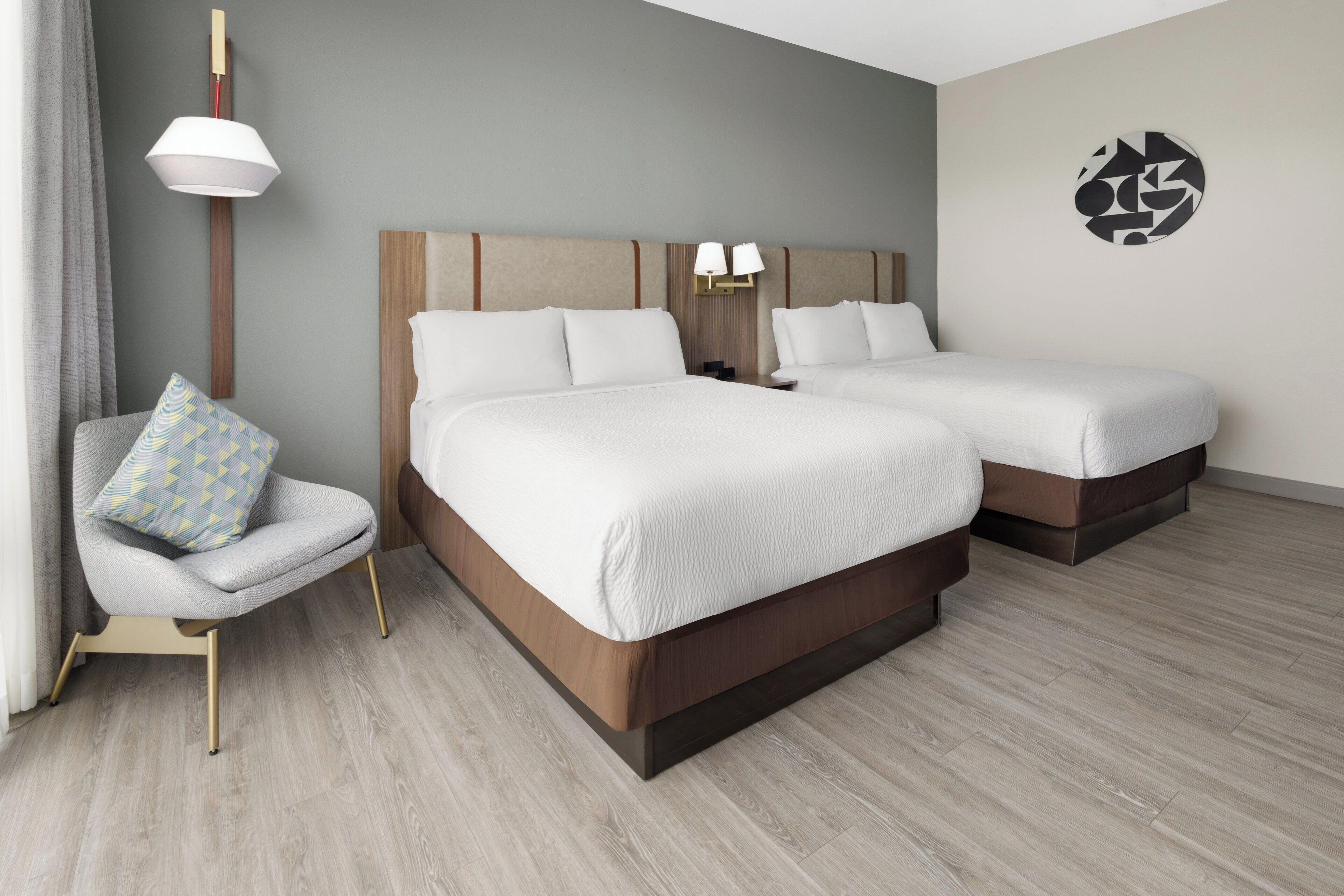 A hotel room with two beds, a chair, a lamp, and a wall art piece.