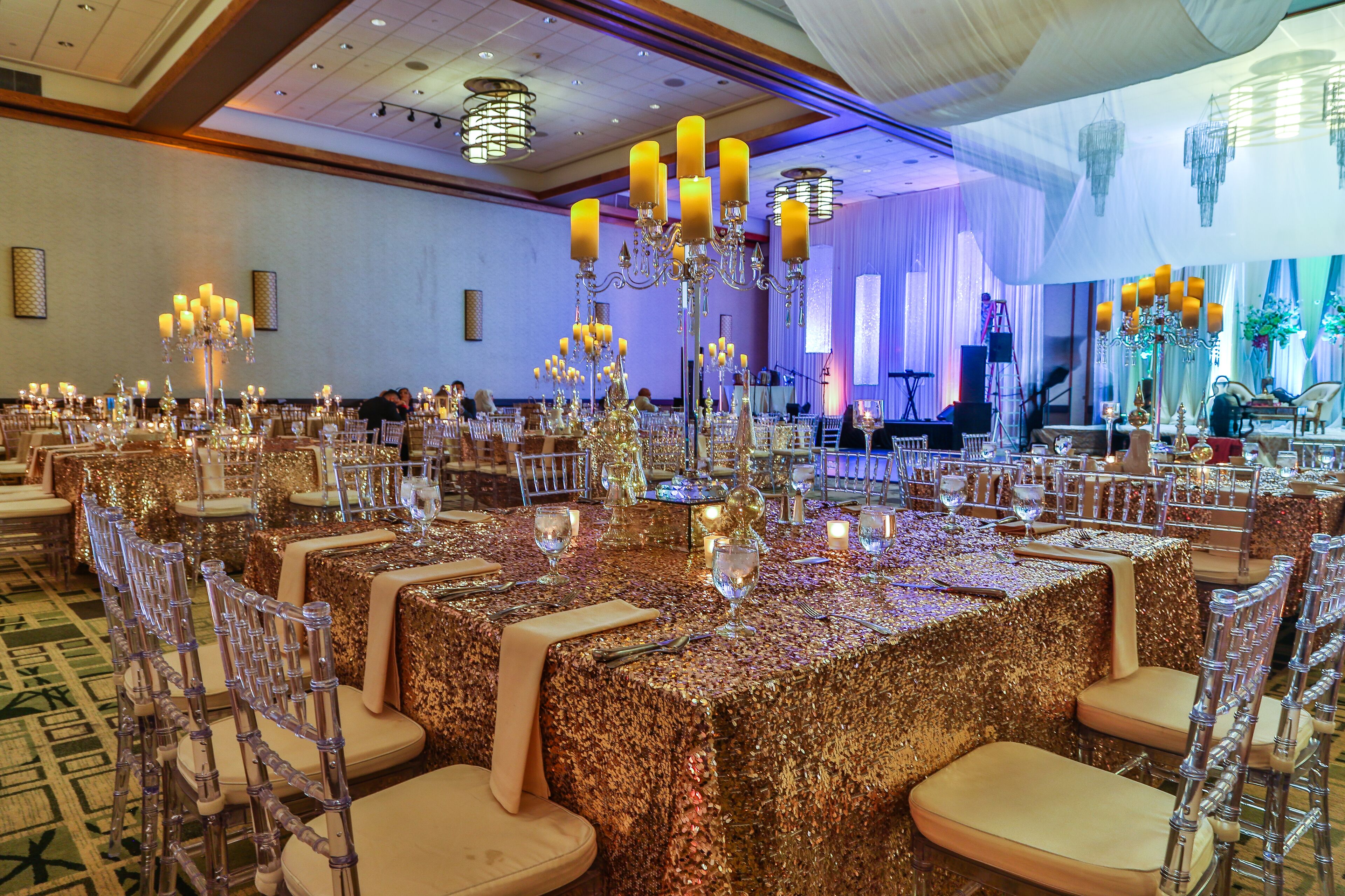 Un salón de banquetes con mesas cubiertas de manteles dorados con lentejuelas, sillas transparentes y candelabros ornamentados.