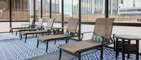 A modern indoor seating area with wicker chairs and a small table, large windows with a city view, and patterned floor mats.