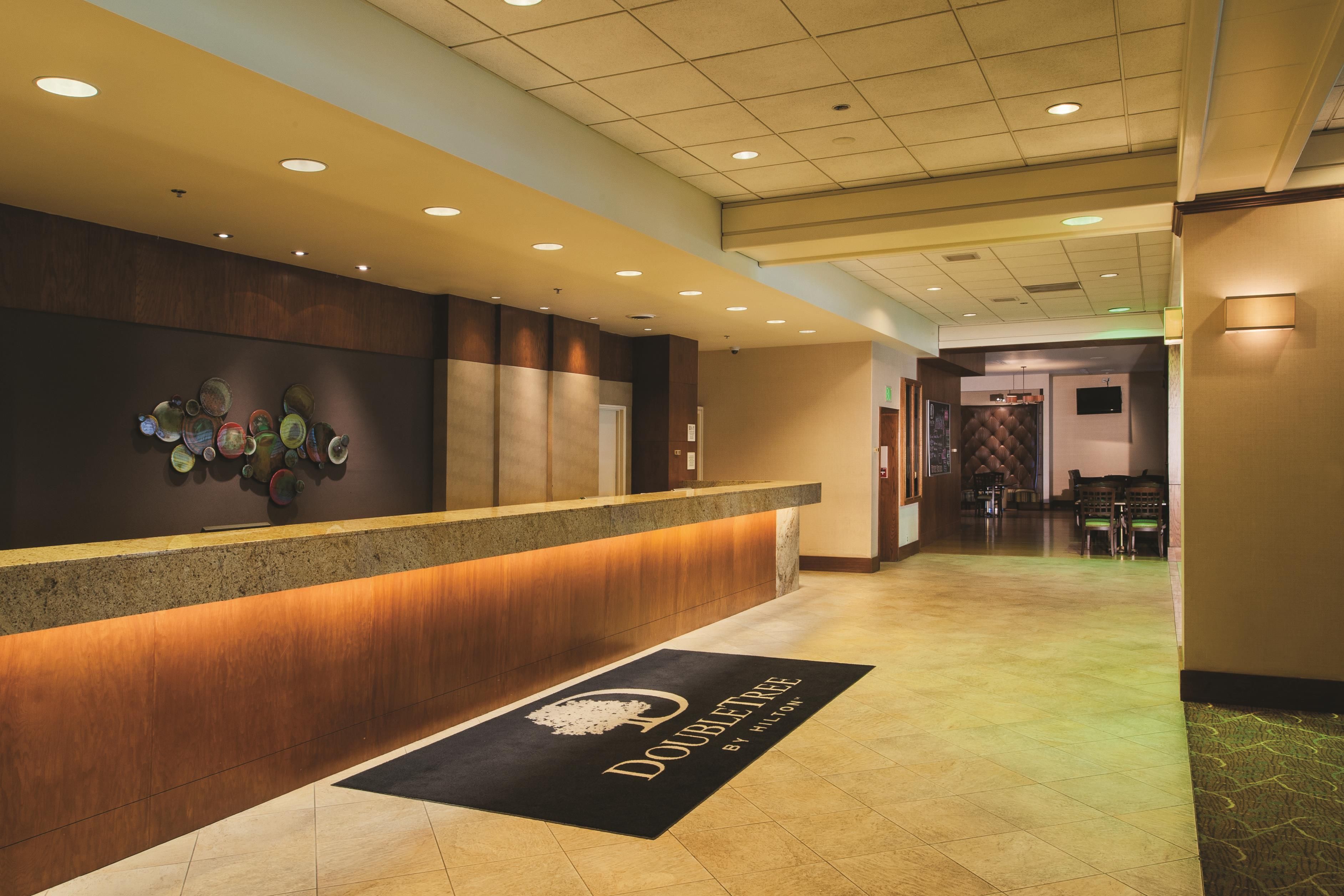A hotel reception area with a wooden reception desk, a wall-mounted art piece, and a doormat with the hotel's name.
