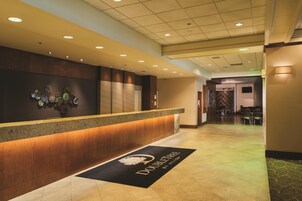 Un hall d’accueil d’hôtel avec un comptoir de réception en bois, une œuvre d’art accrochée au mur et un paillasson portant le nom de l’hôtel.
