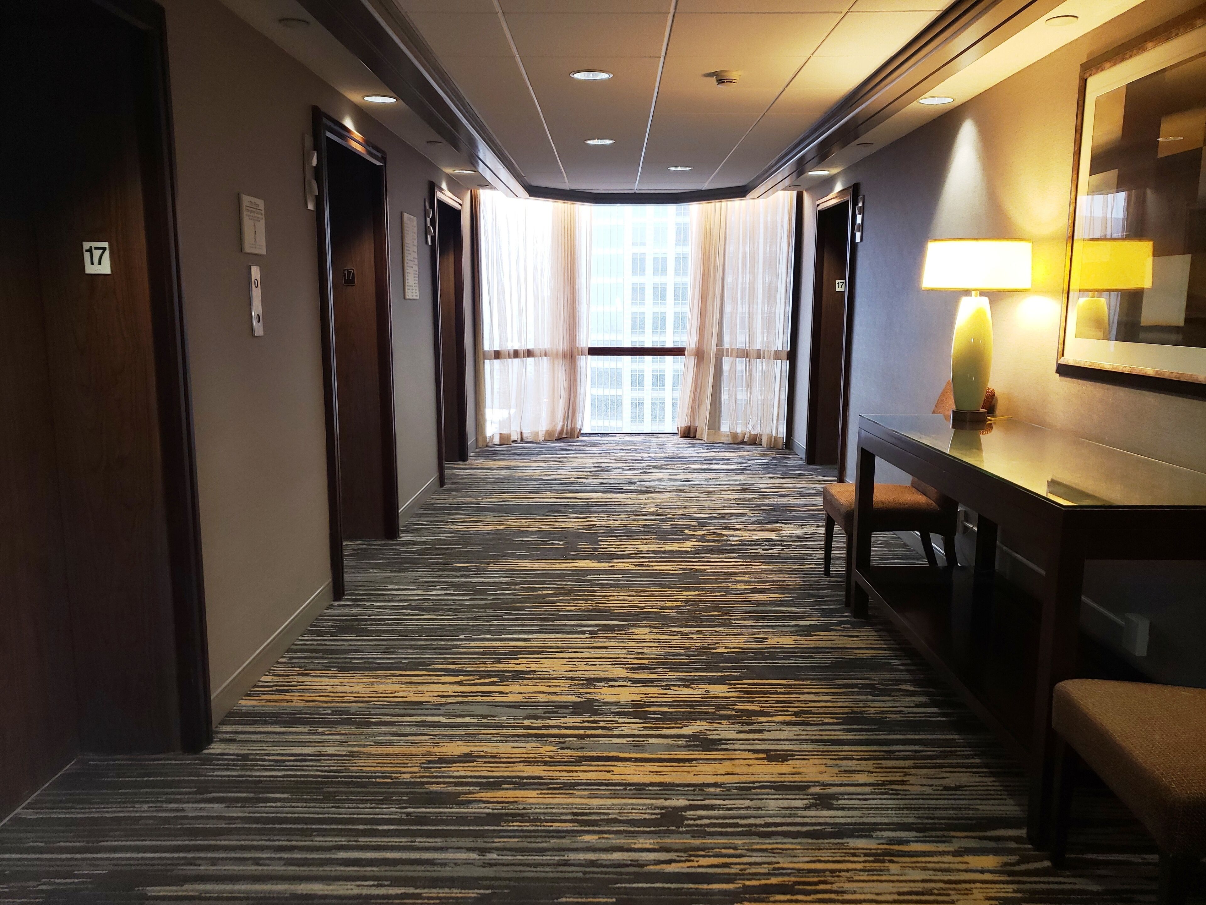 A hotel corridor with a patterned carpet, wooden doors, and a bench with a lamp.