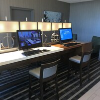 A hotel workspace with two desks, each with a computer monitor, keyboard, and mouse. There are two chairs at each desk. The room has a patterned carpet and a wall with a textured design.