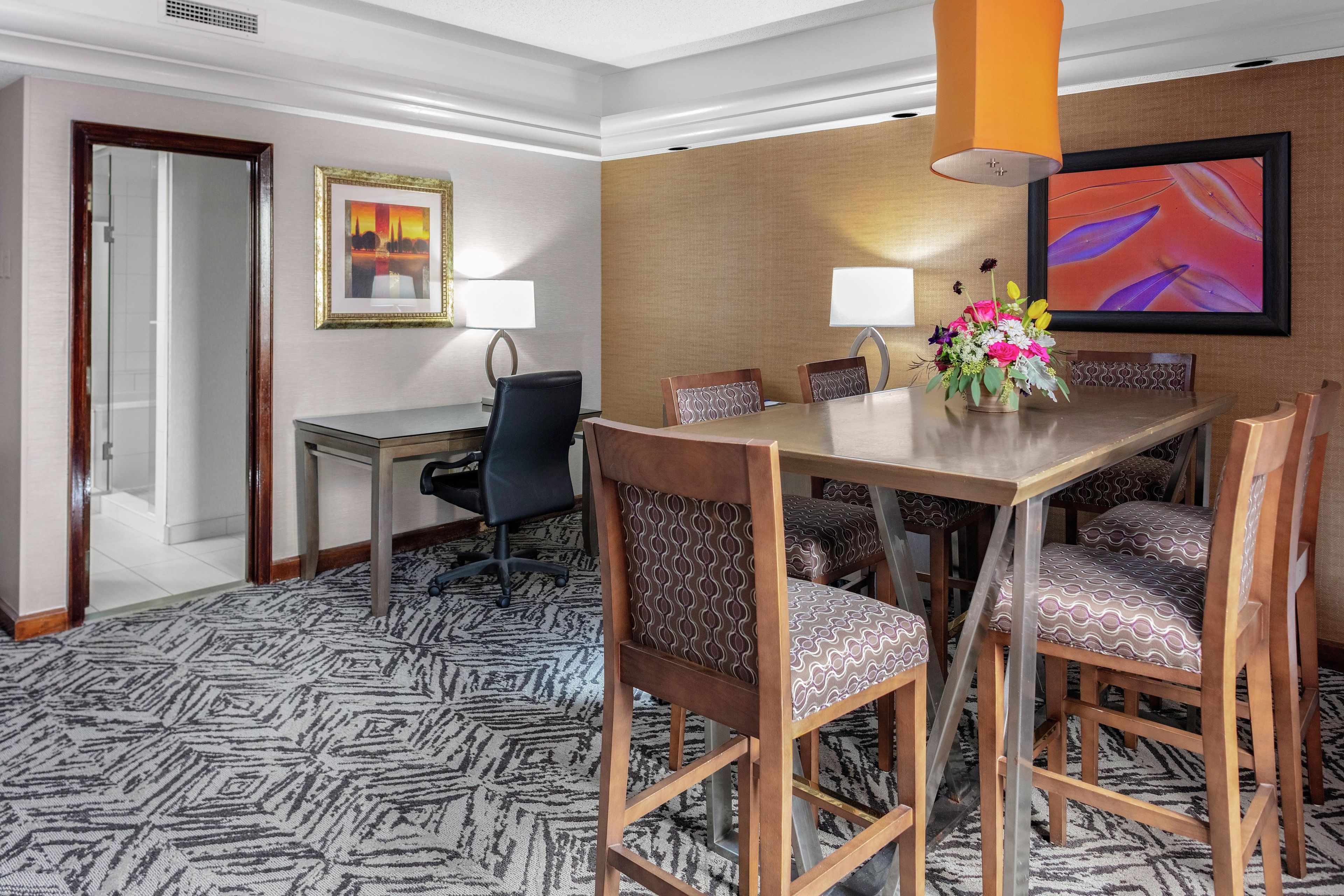 Habitación de hotel con una mesa de comedor, sillas, una lámpara, un cuadro enmarcado y piso alfombrado.