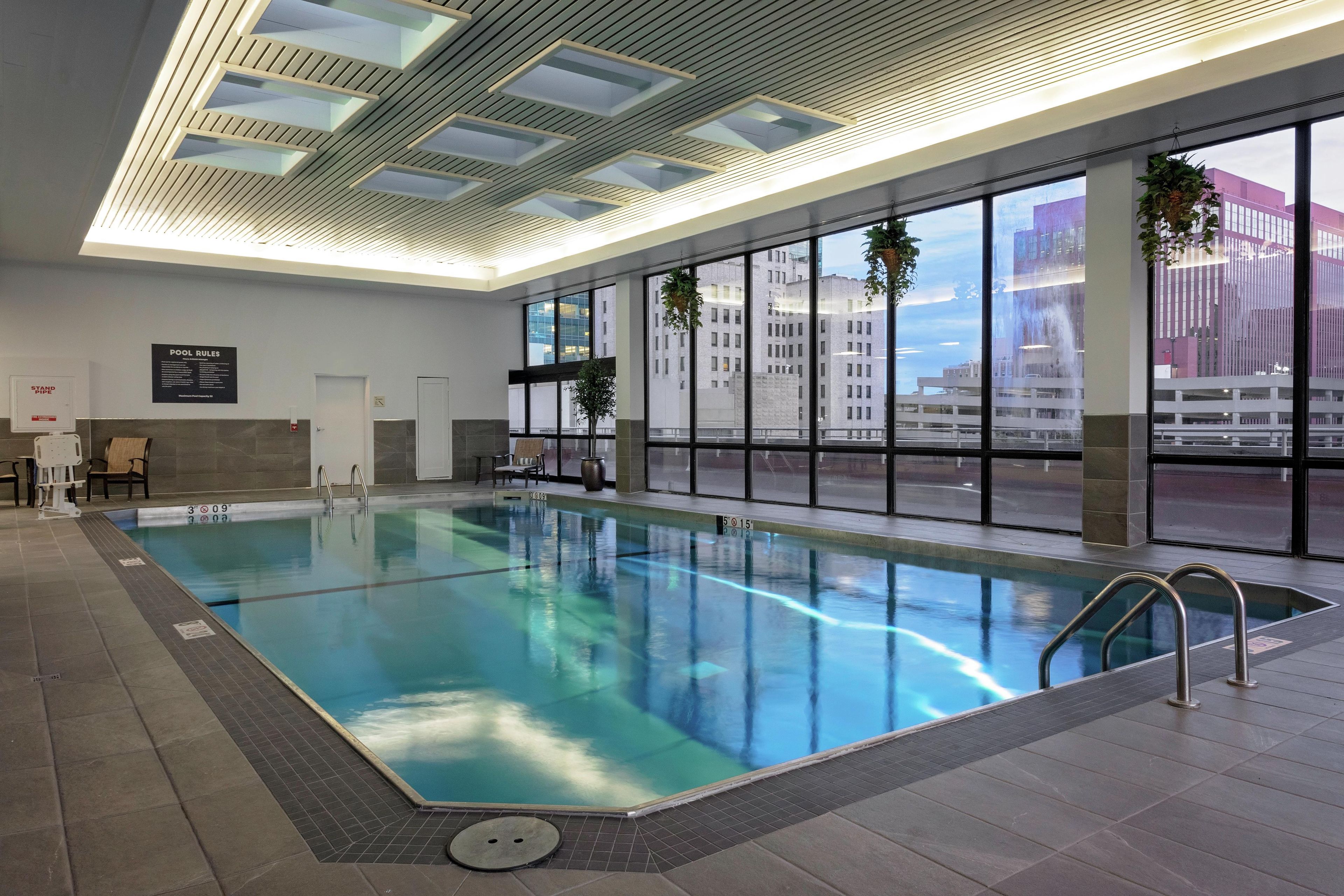 Una piscina interior con agua cristalina, suelo de baldosas y vistas a edificios a través de amplias ventanas.