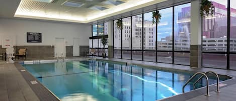 An indoor swimming pool with clear water, a tiled floor, and a view of buildings through large windows.