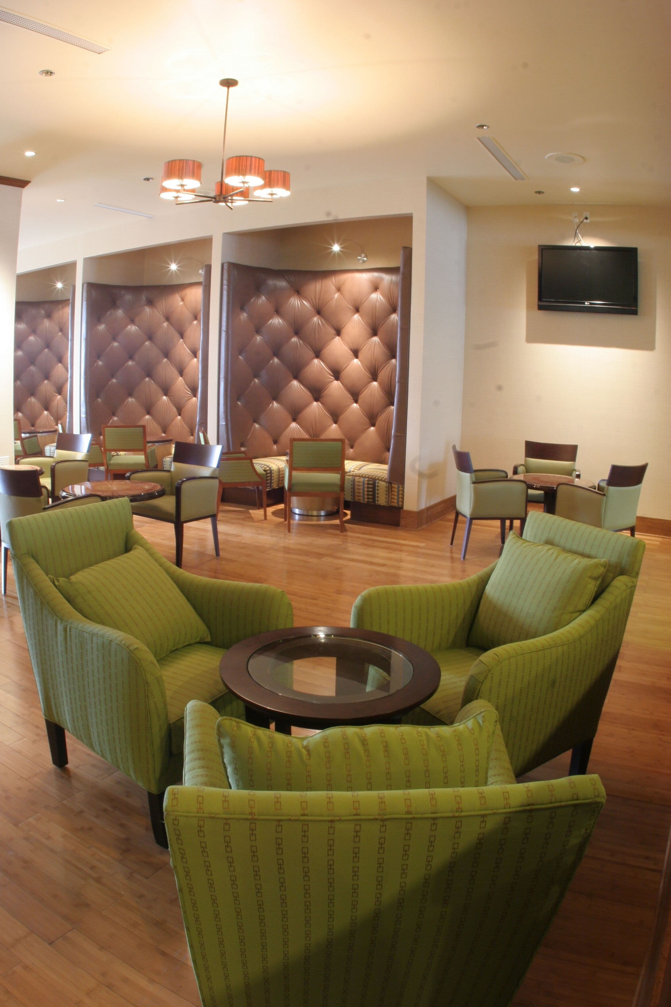 Un hall d'hôtel avec des fauteuils à revêtement vert, une table ronde, un mur en cuir capitonné et un téléviseur fixé au mur.