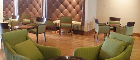 Un hall d'hôtel avec des fauteuils à revêtement vert, une table ronde, un mur en cuir capitonné et un téléviseur fixé au mur.