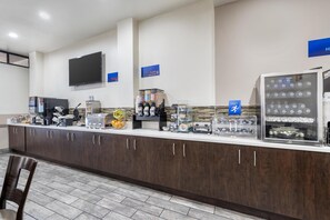 Zona de desayuno de hotel con un mostrador de autoservicio, máquinas de café y un refrigerador.