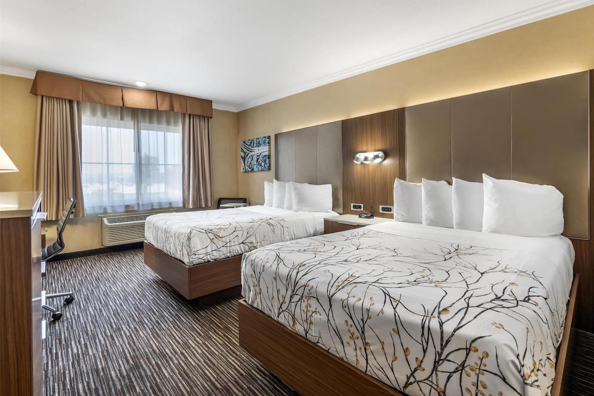 A hotel room with two beds, a desk, a chair, a window with curtains, and a wall-mounted light fixture.