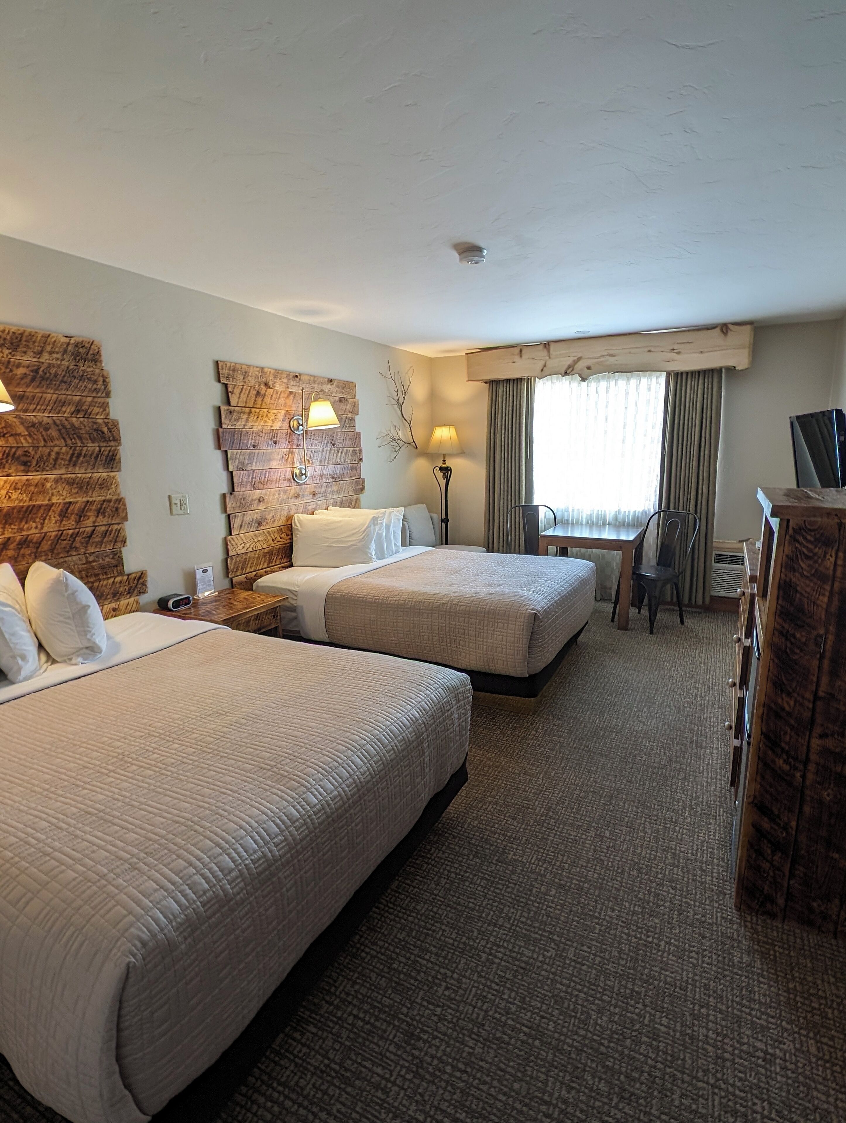 Quarto de hotel com duas camas, uma mesa, uma cadeira, uma luminária e uma televisão.