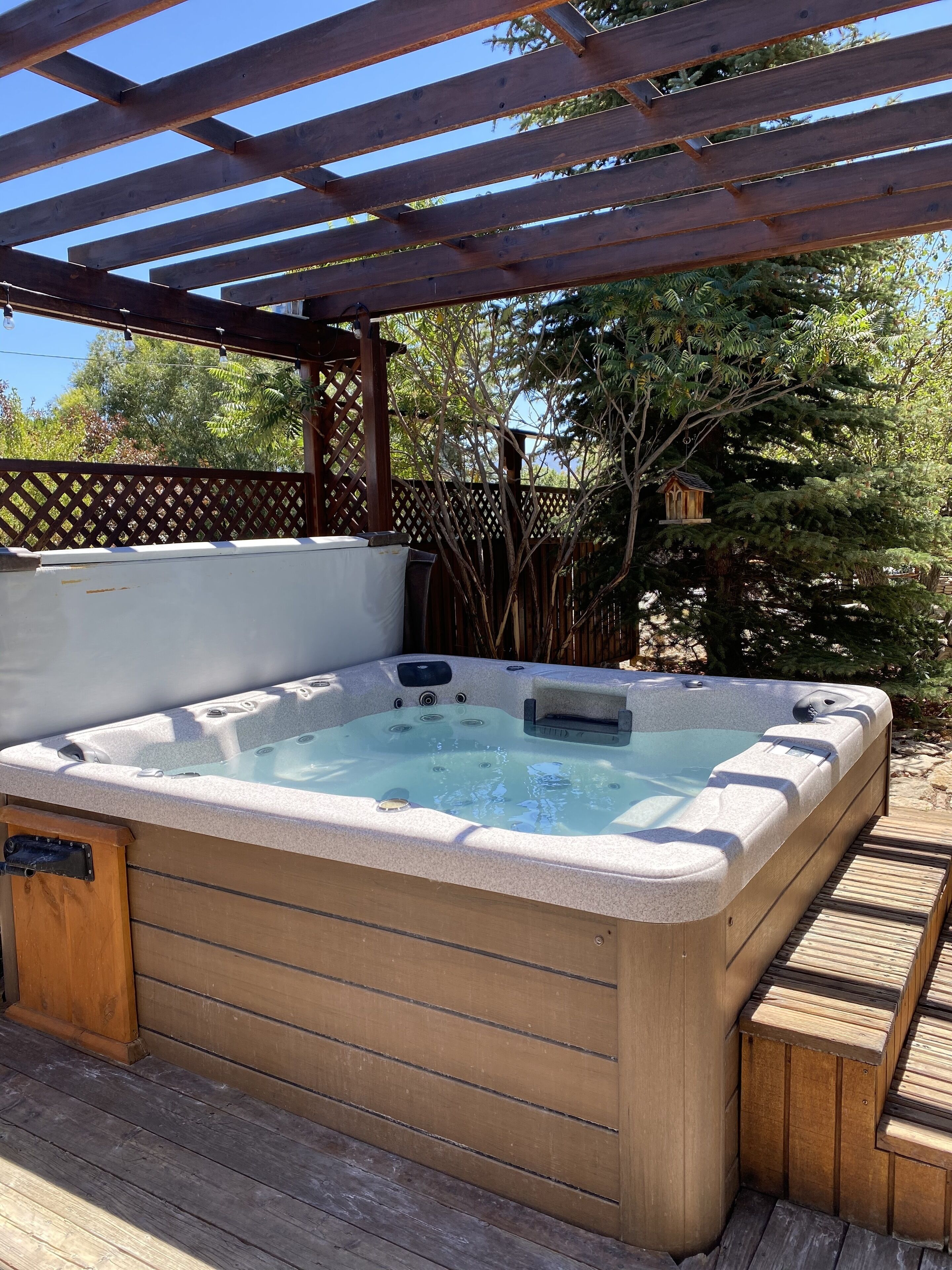 Un bain à remous sous une pergola en bois, avec une terrasse en bois et une clôture en treillis en arrière-plan.