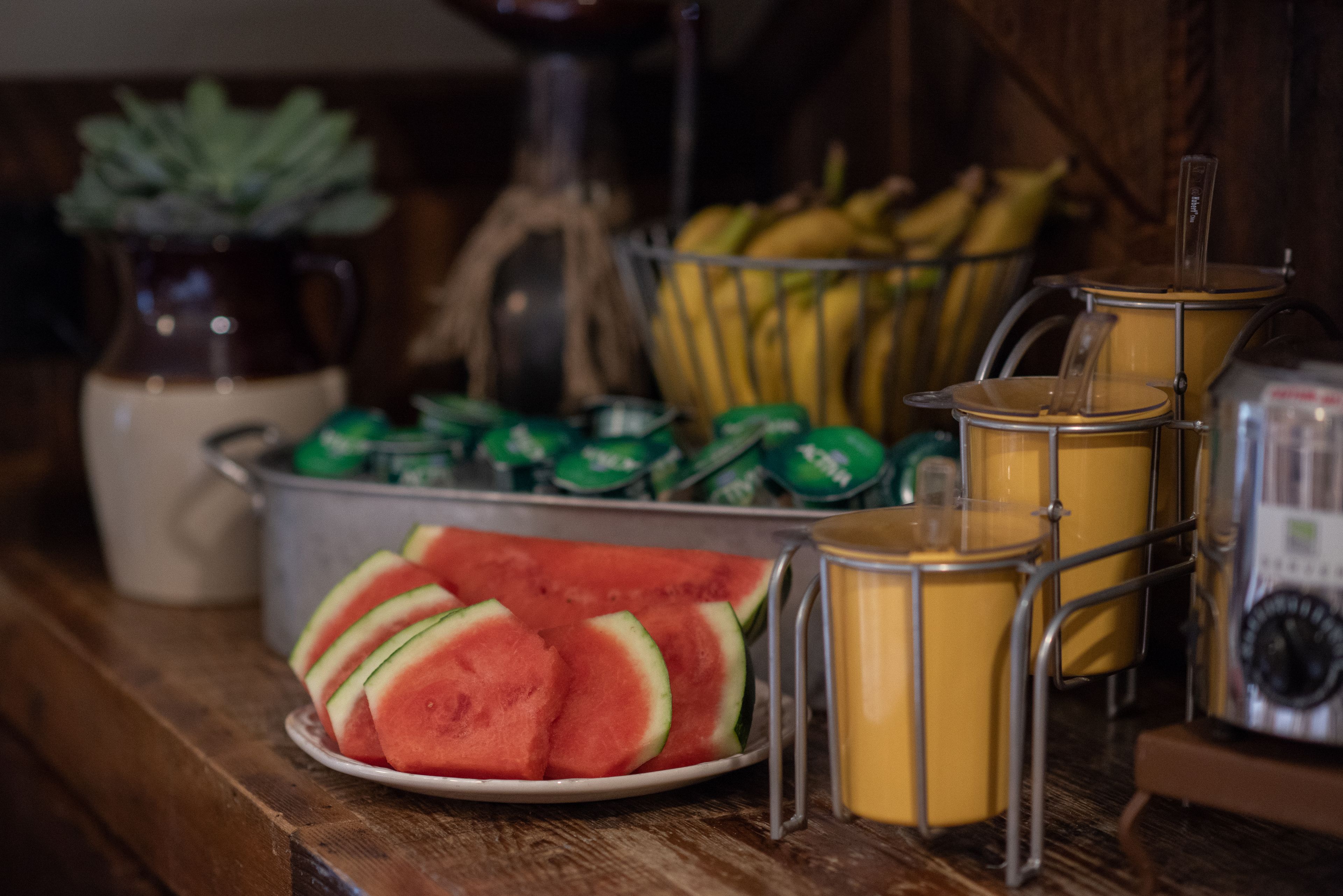 A table with a plate of sliced watermelon, a metal basket with bananas, and a metal container with green cans.