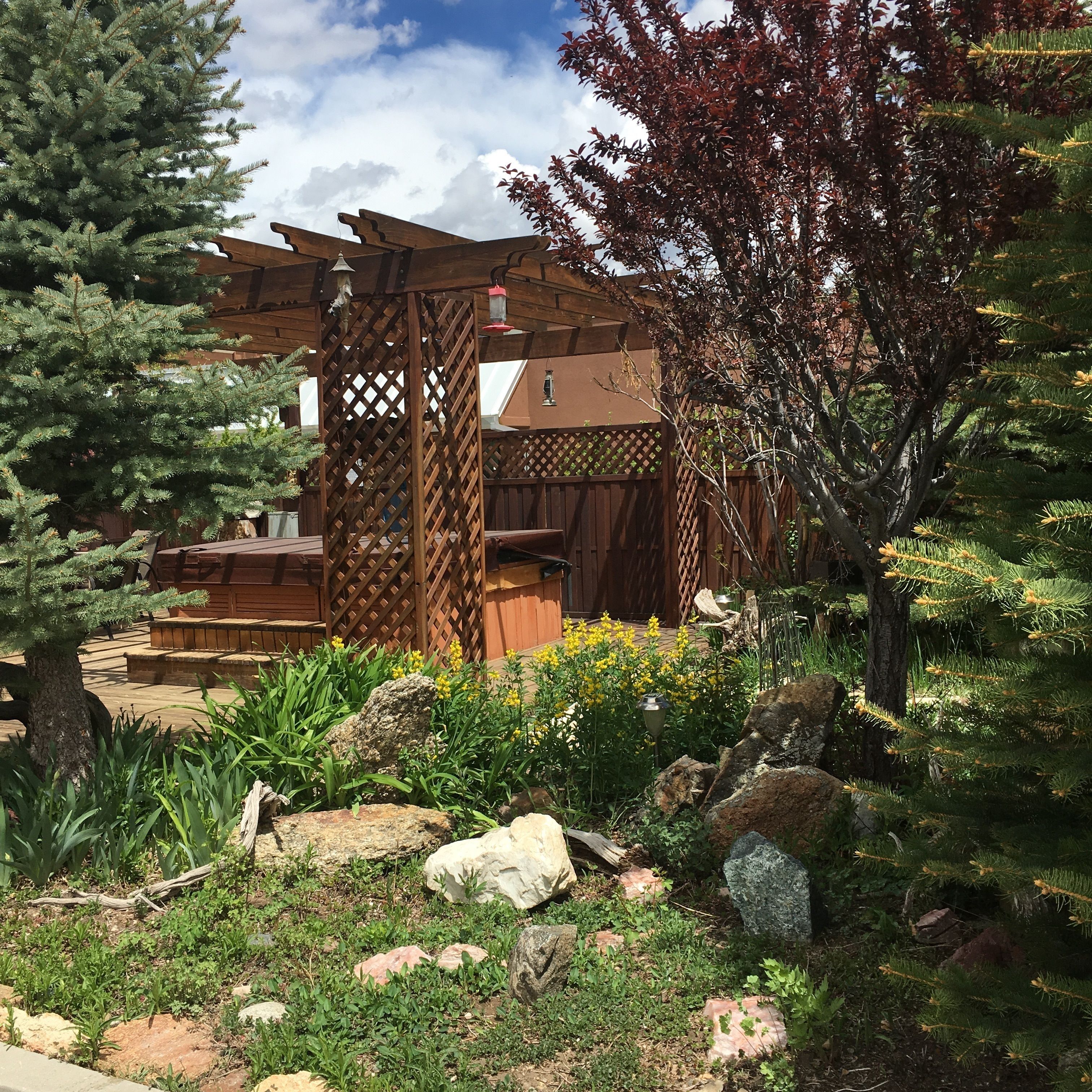 A wooden pergola structure with a lattice design, surrounded by a garden with various plants and rocks.