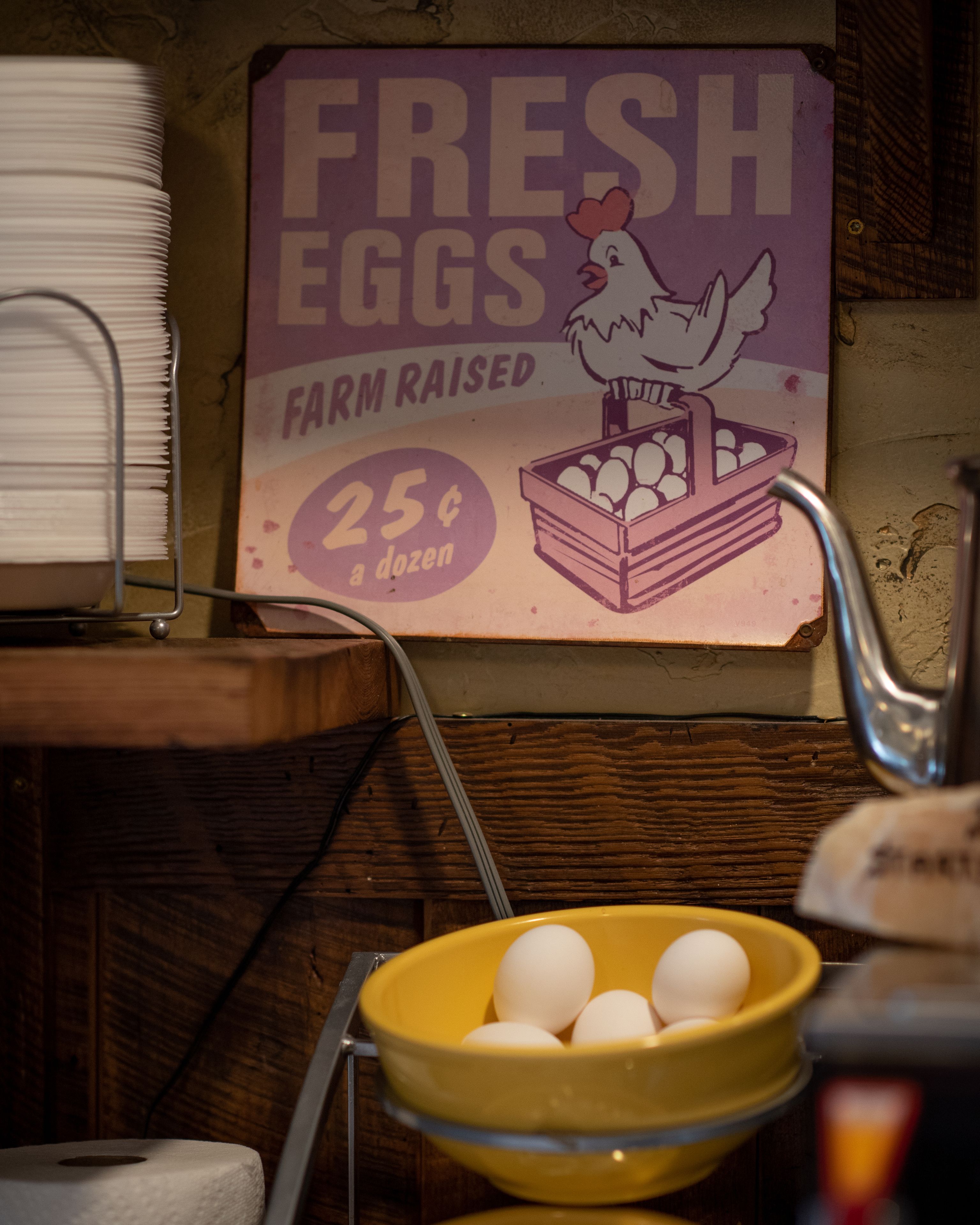 Letrero en la pared que anuncia huevos frescos a 25 centavos la docena, un tazón amarillo con huevos y un estante metálico con toallas de papel.