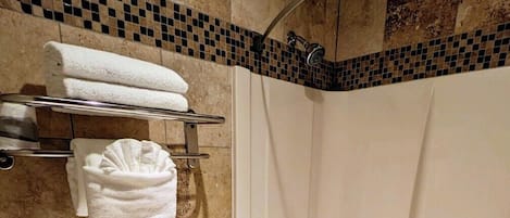 A bathroom with a toilet, bathtub, and shower. There are towels on a shelf and a trash bin on the floor.