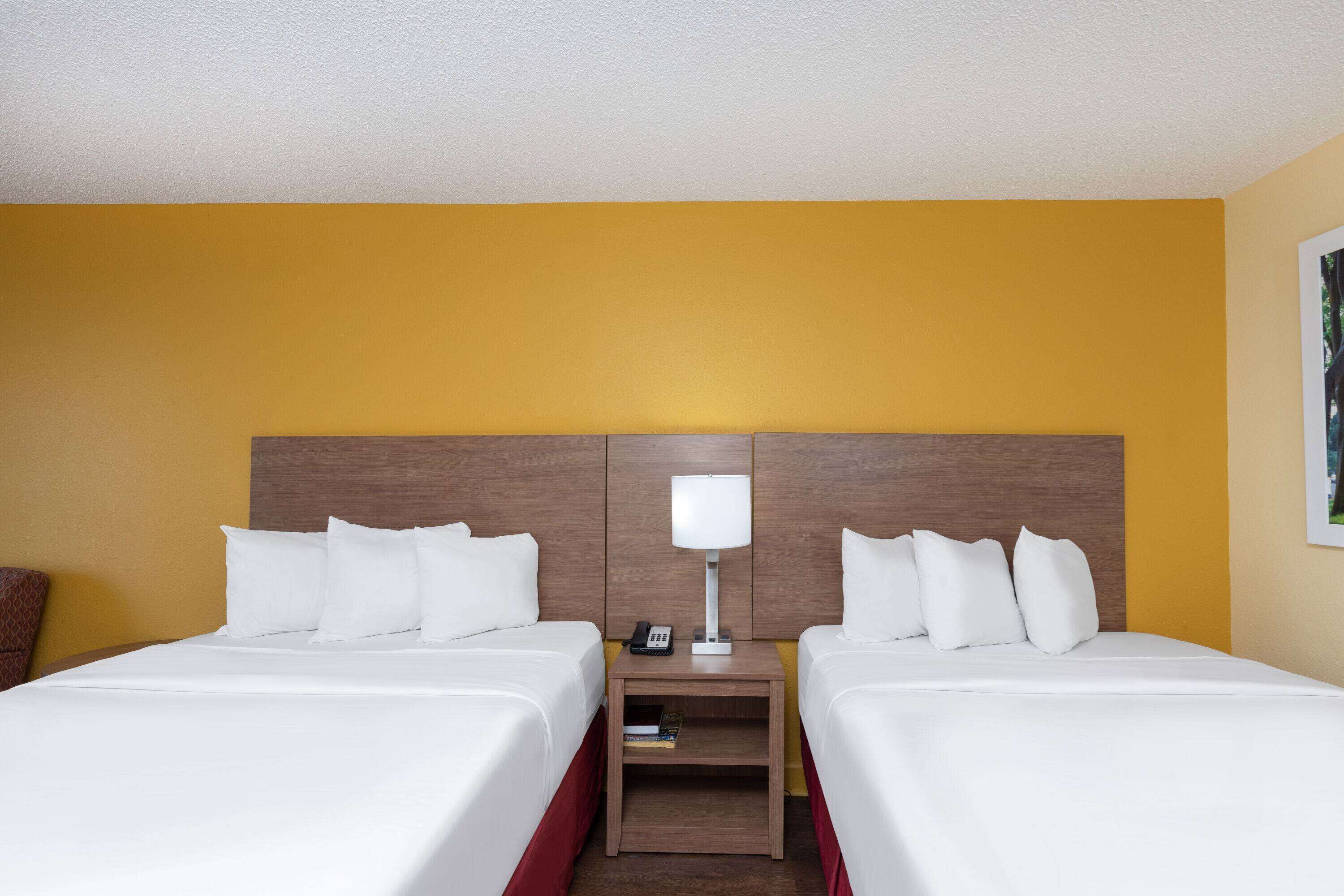 A hotel room with two beds, a nightstand with a lamp, and a small table with a book.