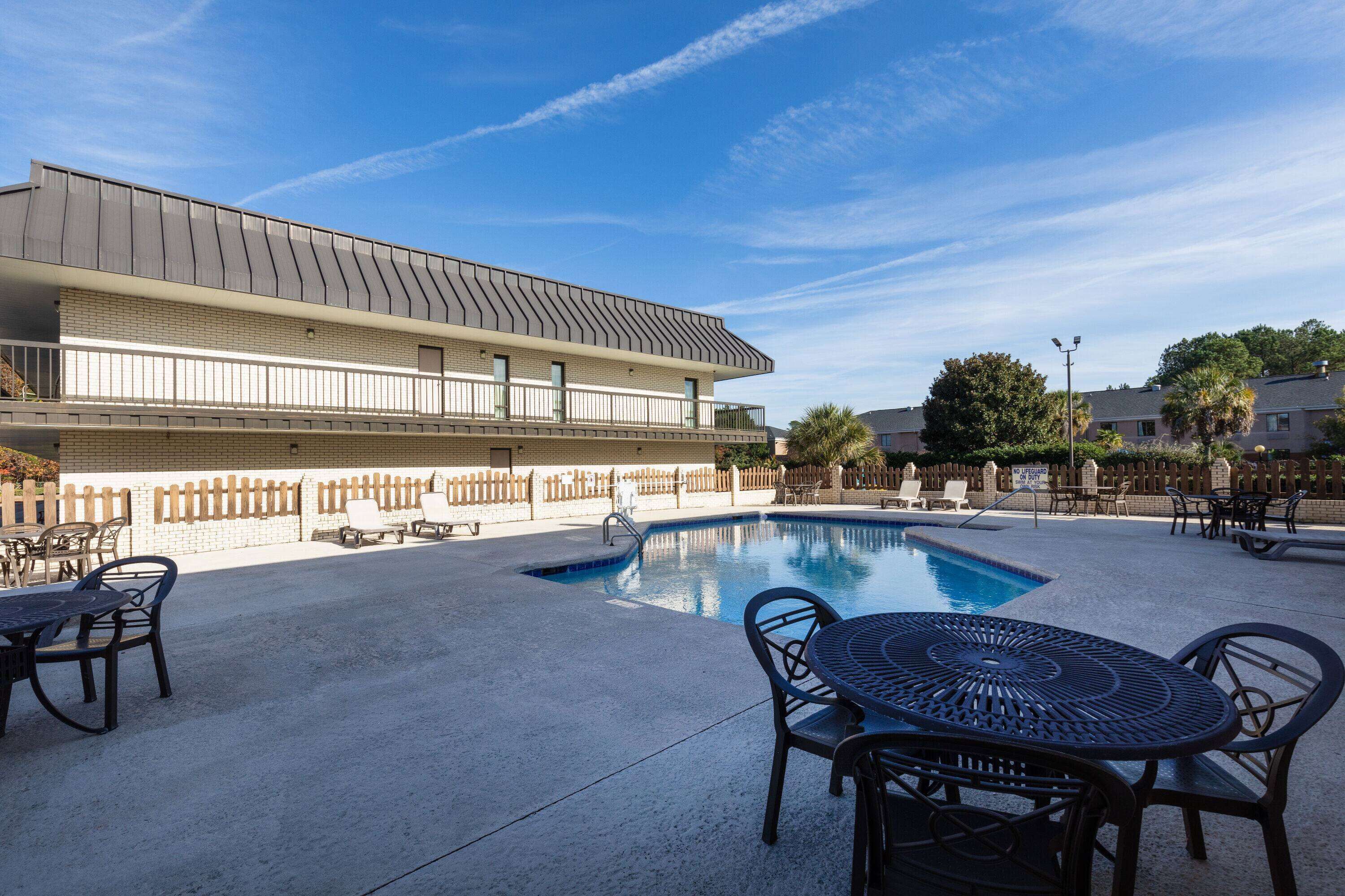 Uma área de piscina com espreguiçadeiras e uma mesa, um edifício com telhado plano e um céu azul e límpido.