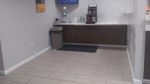 A small, clean room with a counter, a trash bin, a coffee machine, and a TV screen displaying a news program. - Motel 6 Carlisle, PA - Cumberland Valley (Carlisle)