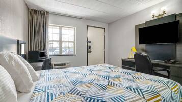 A hotel room with a large bed, a desk with a computer, a TV mounted on the wall, and a window with curtains.