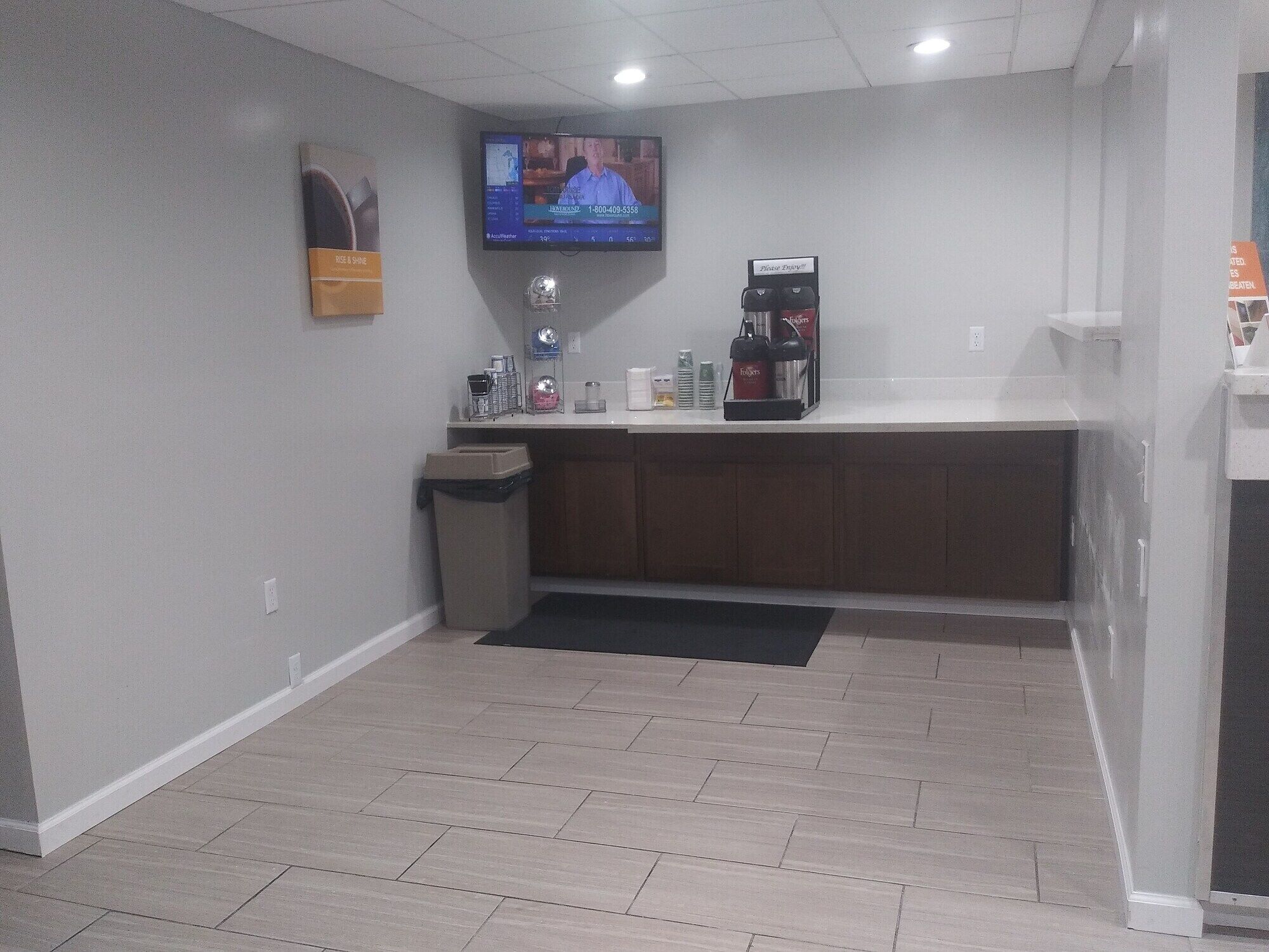A small, clean room with a counter, a trash bin, a coffee machine, and a television mounted on the wall.