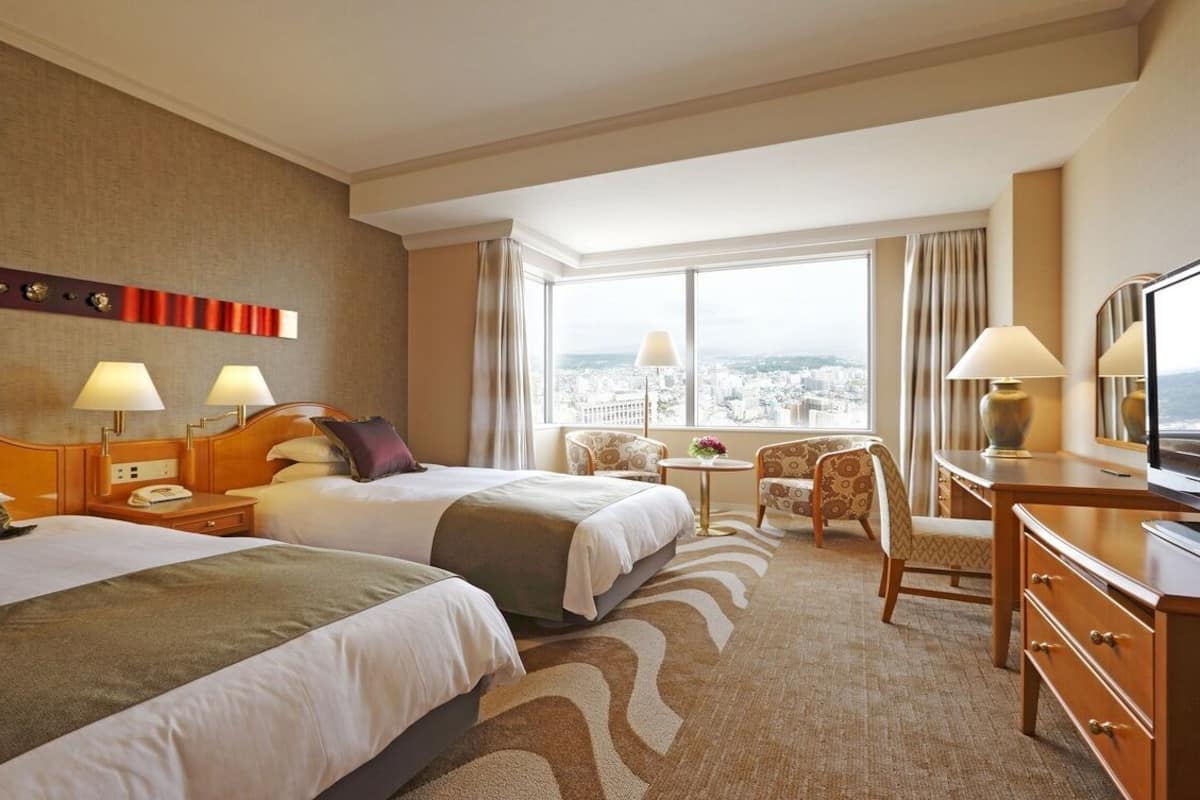 A hotel room with two beds, a desk, a chair, and a large window offering a city view.