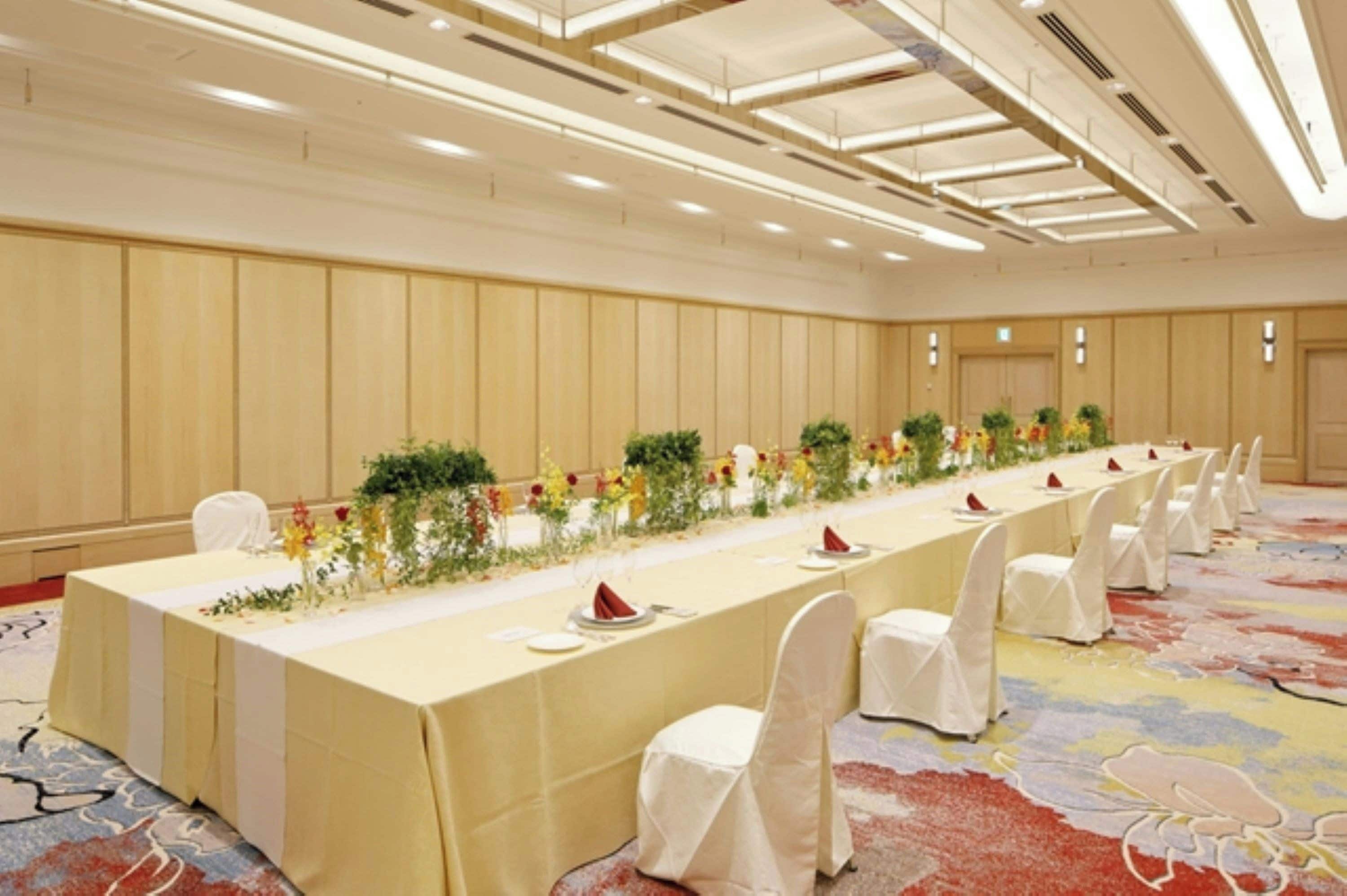 A long banquet table with white chairs, set with plates and napkins, in a room with wooden paneling and a patterned carpet.