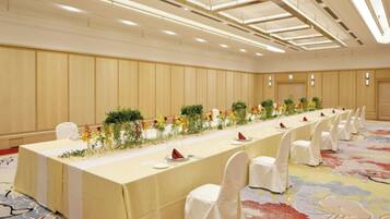 A long banquet table with white chairs, set with plates and napkins, in a room with wooden paneling and a patterned carpet.