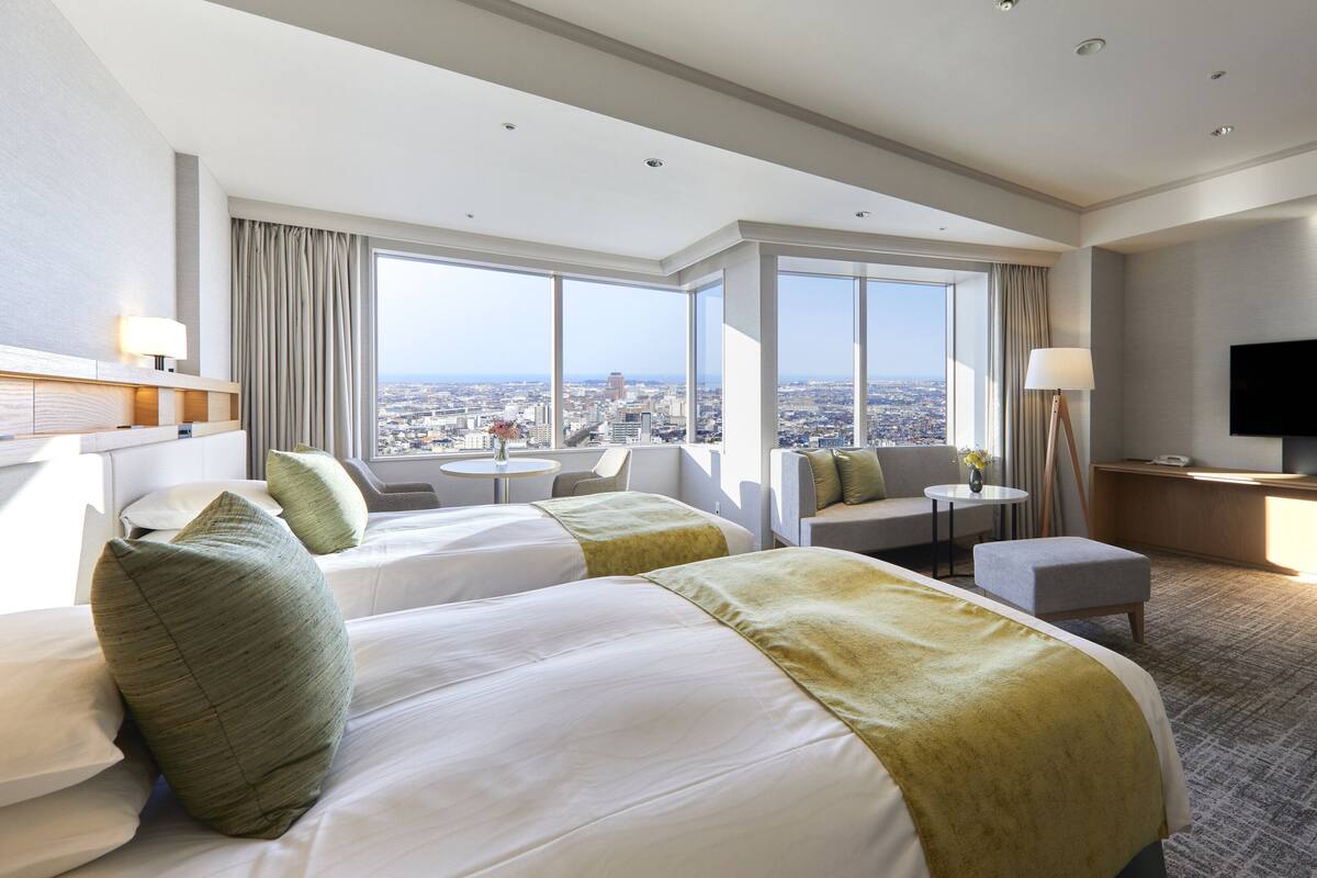 A hotel room with two beds, a desk, a TV, and a view of the city.