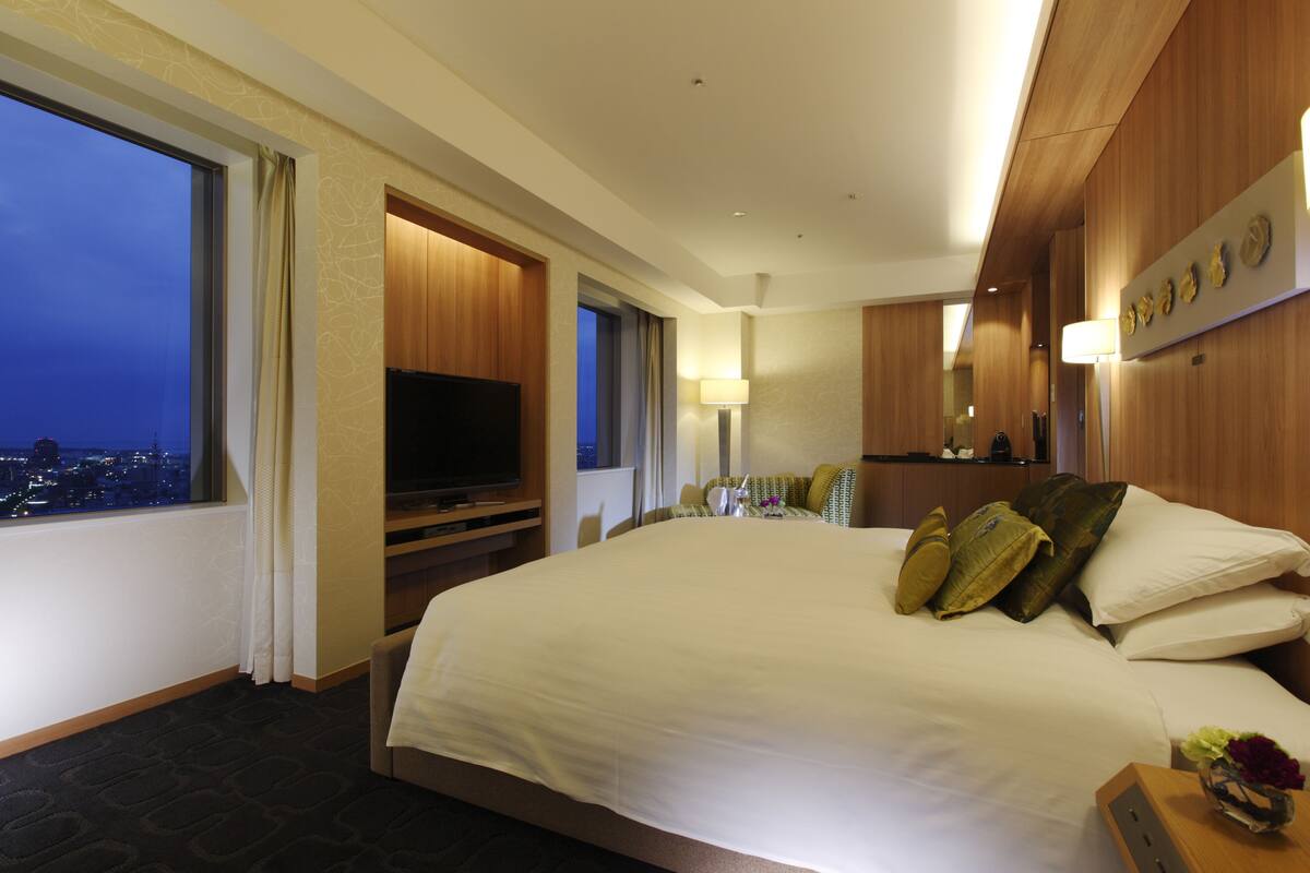 A hotel room with a large bed, a television, a desk, and a view of the city at night.
