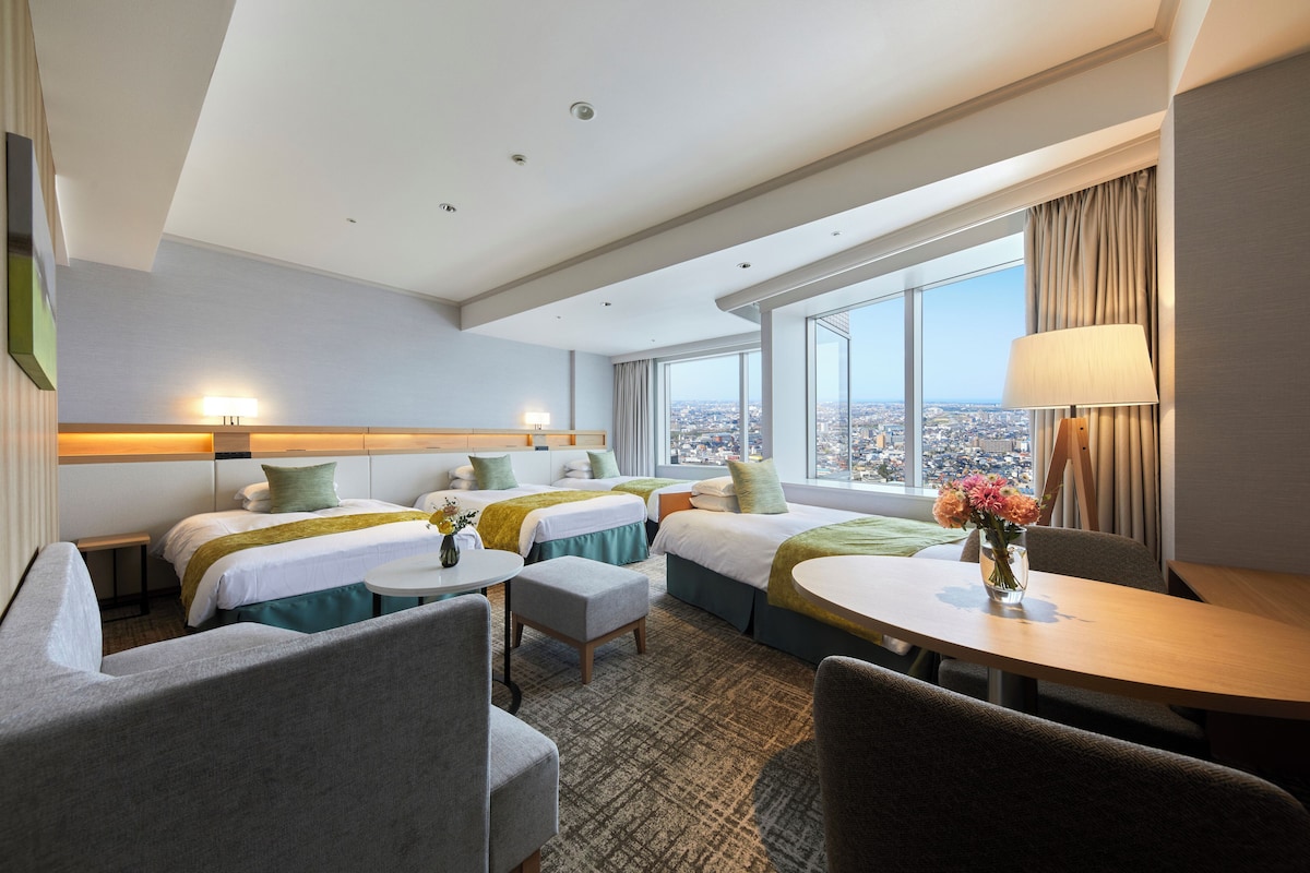 Habitación de hotel con dos camas, una mesa de comedor, un pequeño rincón de estar y un ventanal con vistas a la ciudad.