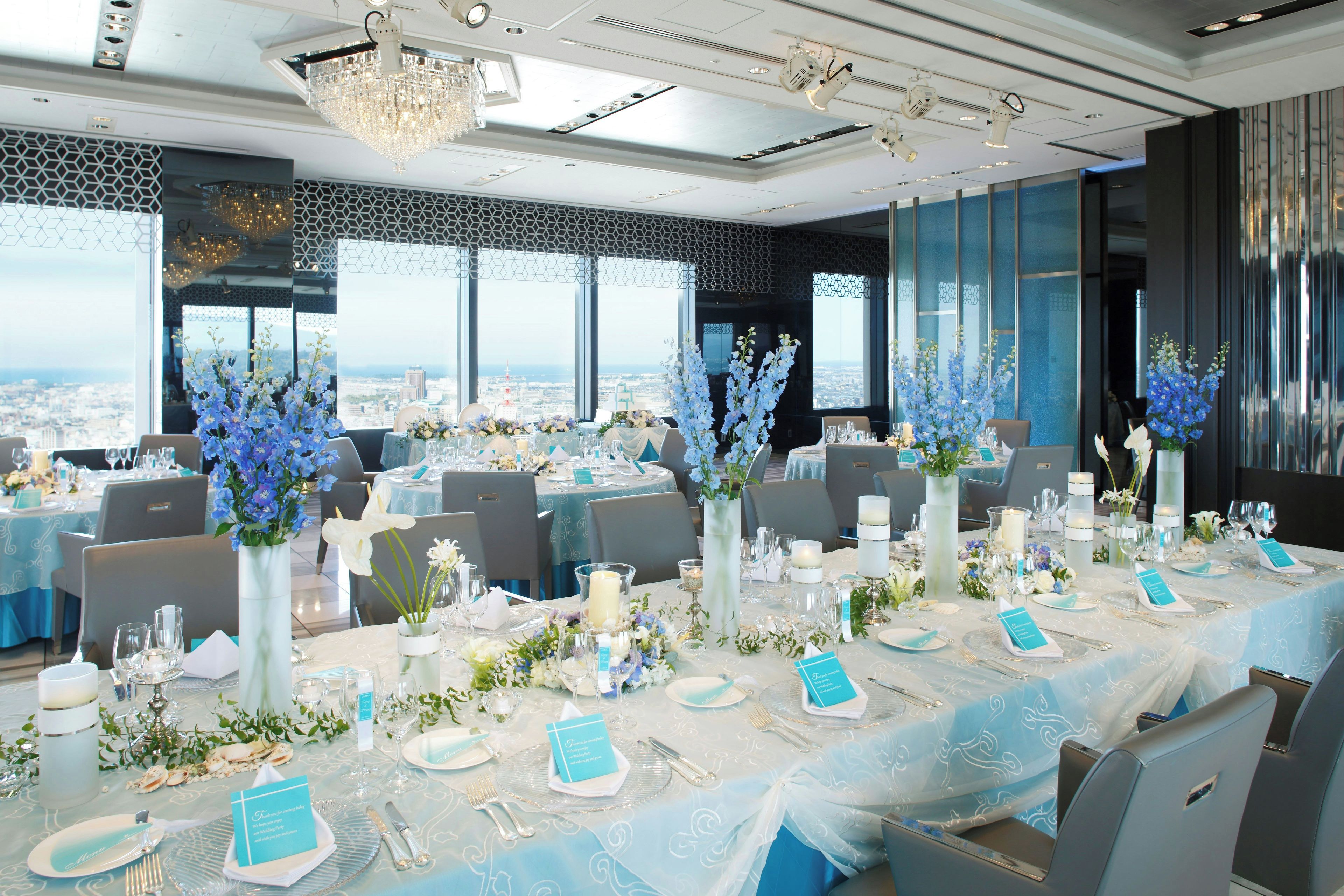 Un salón de banquetes con mesas redondas dispuestas para un evento formal, con centros de mesa florales y vistas a la ciudad.