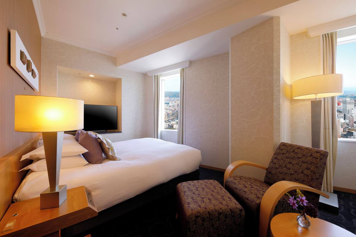 A hotel room with a bed, a TV, a desk with a lamp, a chair, and a view of the city through the window.