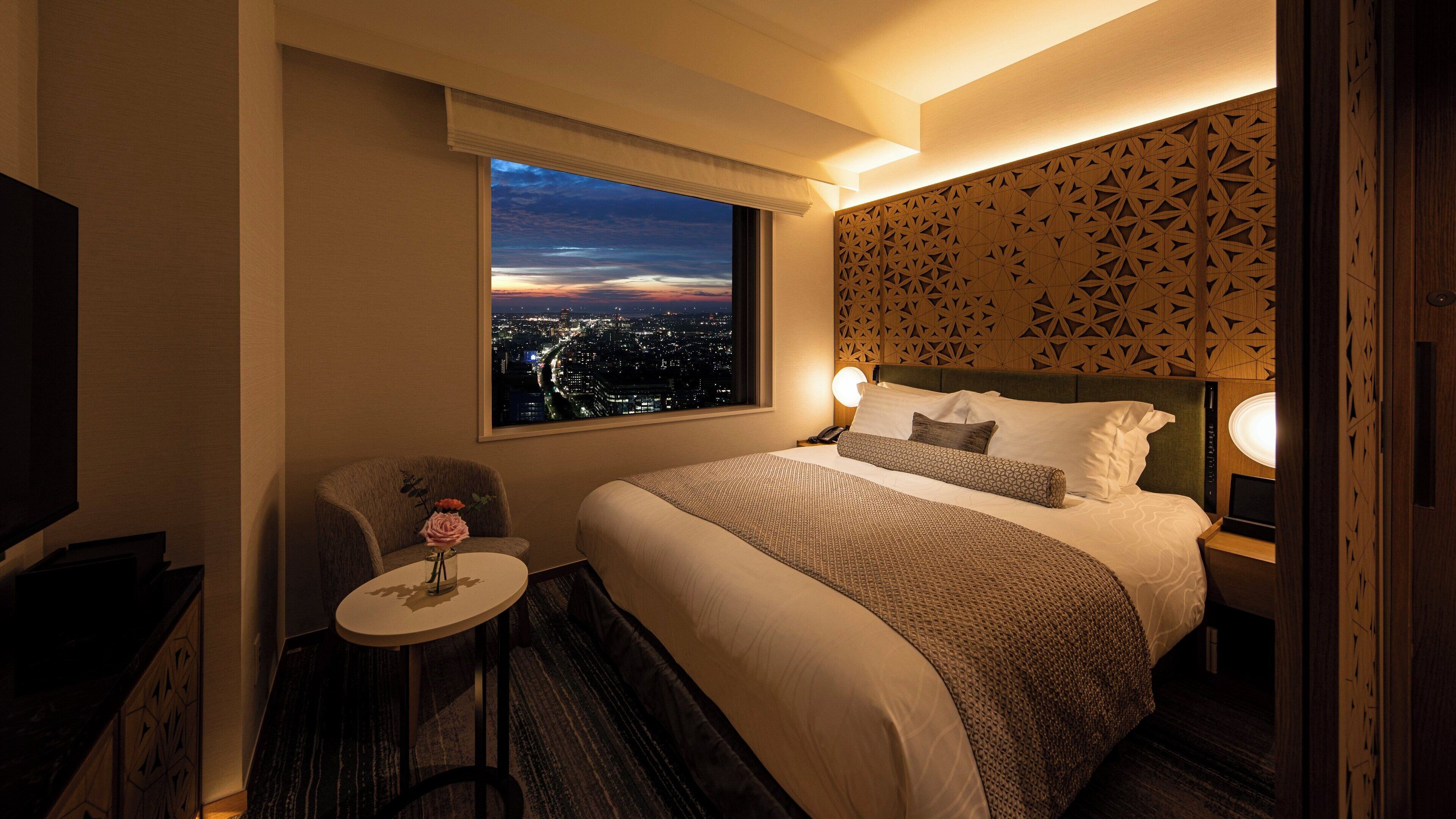 Habitación de hotel con una cama grande, una silla, una mesita con un florero y vistas a la ciudad a través de la ventana.