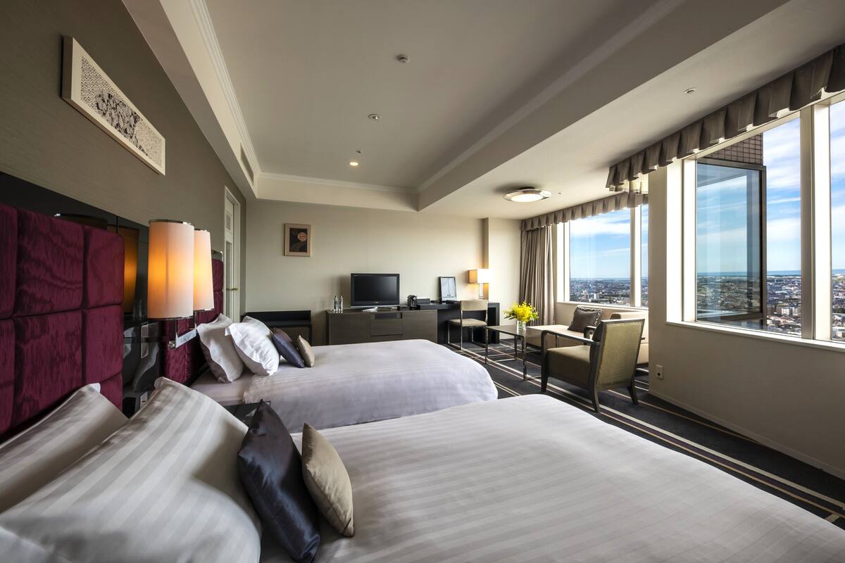 A hotel room with two beds, a desk, a TV, and a large window with a city view.
