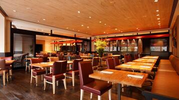 Un restaurante moderno con mesas y sillas de madera, un techo alto con iluminación empotrada y un amplio ventanal que proporciona luz natural.