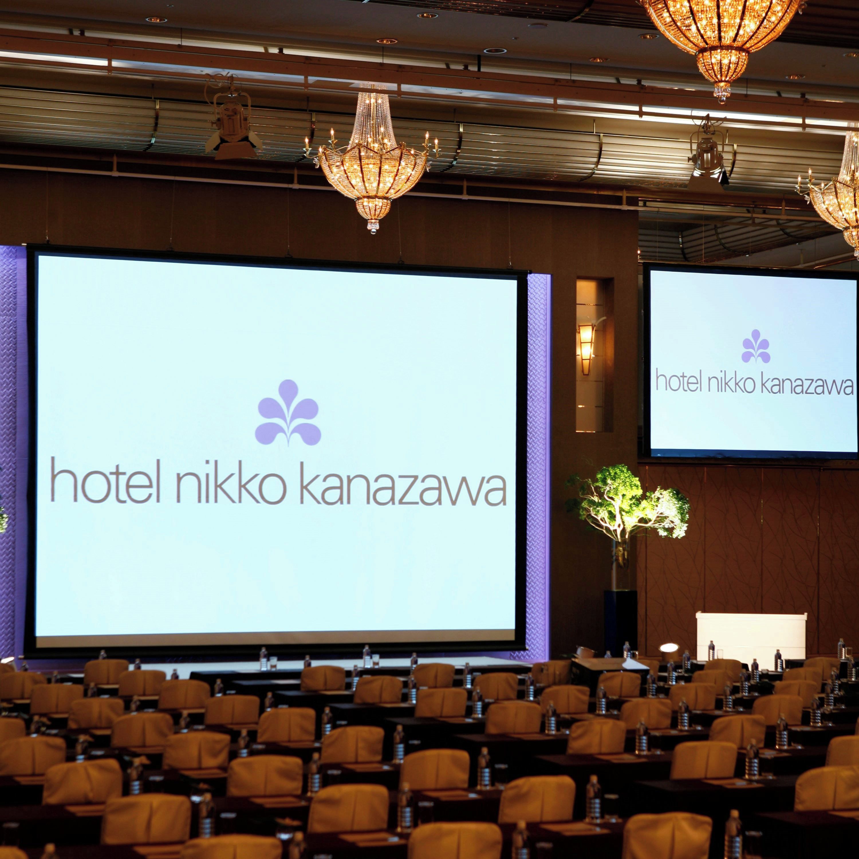 A conference room with a large screen displaying the hotel name, rows of chairs, and chandeliers.