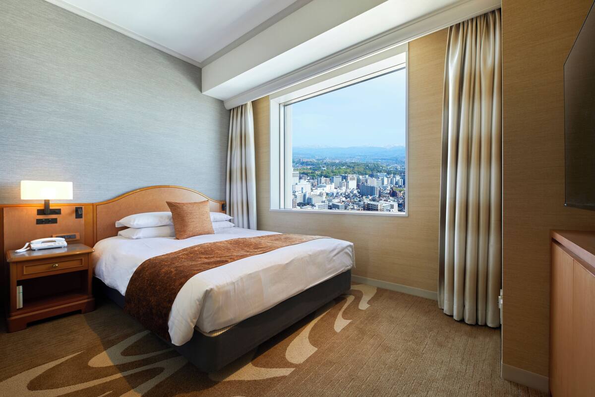 A hotel room with a large bed, a nightstand, a lamp, and a view of a cityscape through a large window.