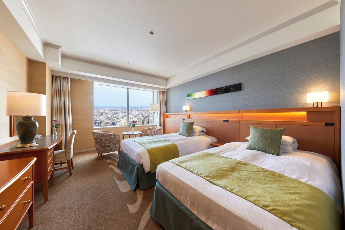 A hotel room with two beds, a desk, a chair, and a view of the city.