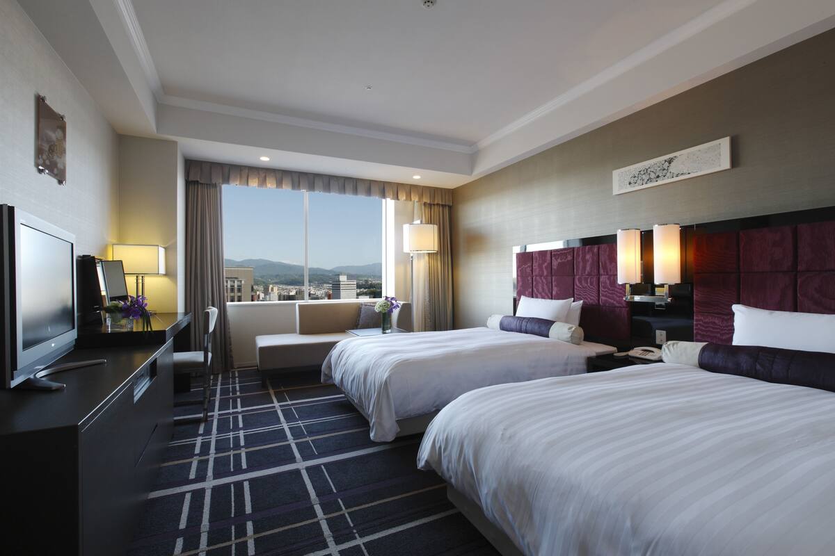 A hotel room with two beds, a TV, a desk, and a view of the city.