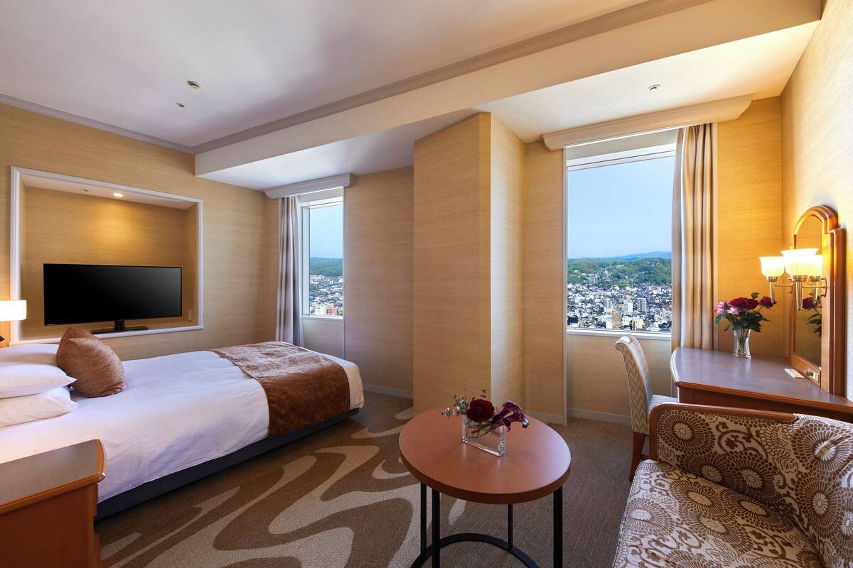 A hotel room with a bed, a TV, a small table with a vase of flowers, a sofa, and a view of a cityscape through large windows.