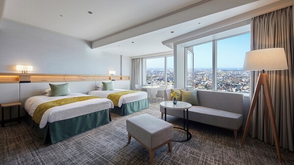 A hotel room with two beds, a sofa, a small table, a lamp, and a view of the city.