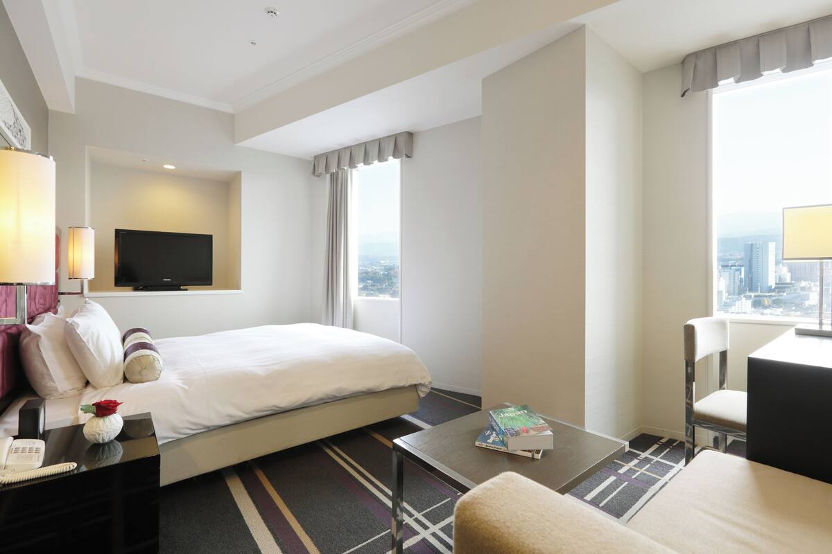 A hotel room with a bed, a desk, a chair, a television, and a city view.