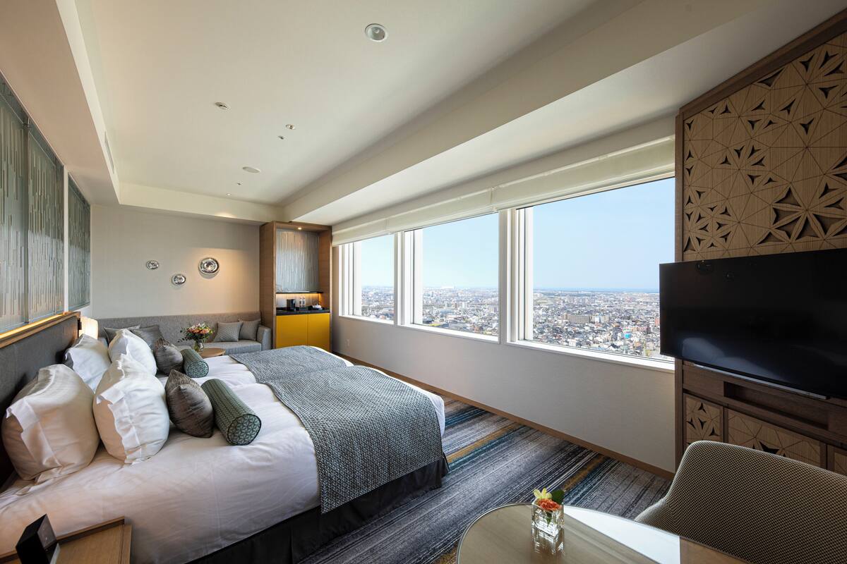 A hotel room with a large bed, a television, a city view, and a window with patterned blinds.