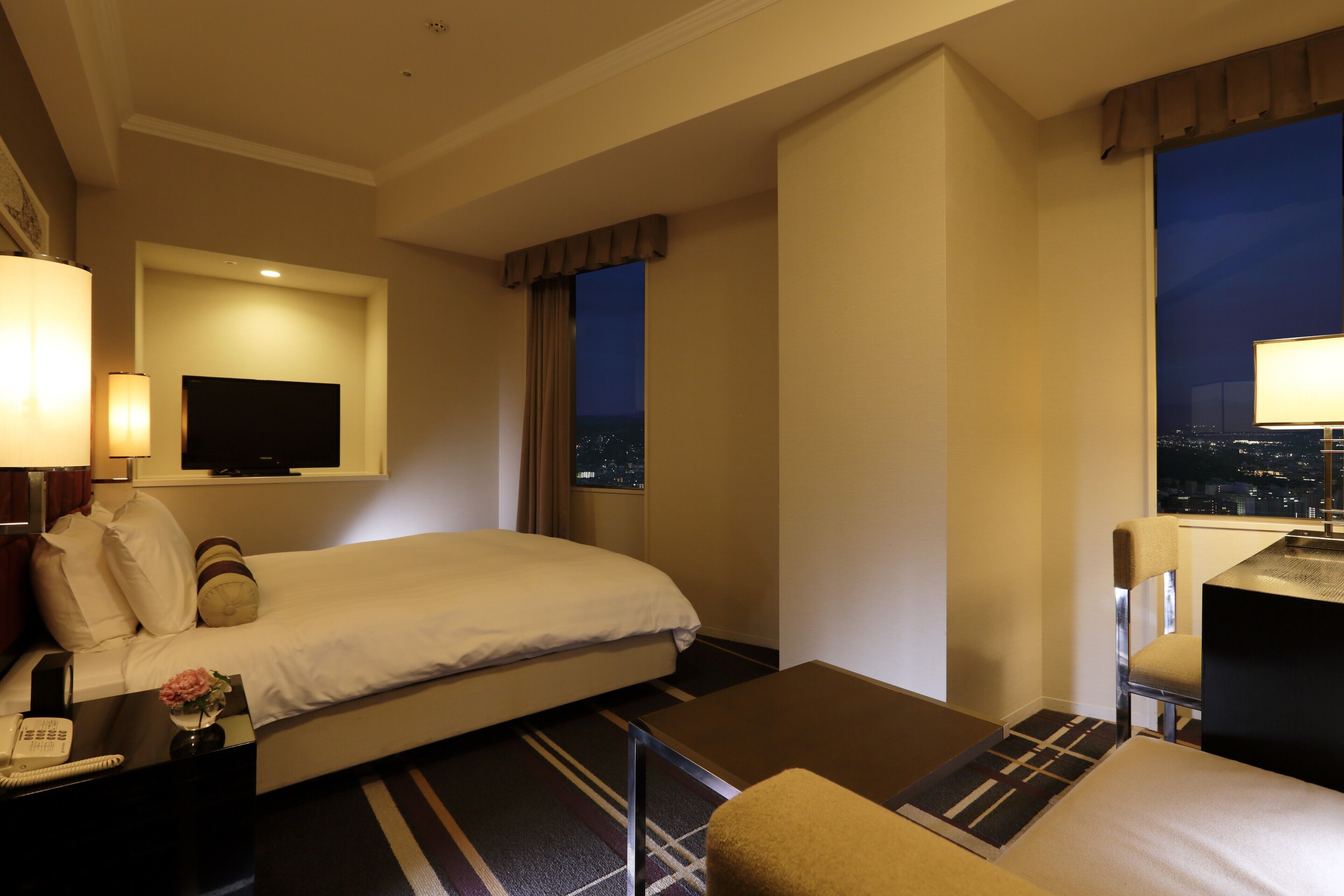 A hotel room with a bed, a desk, a chair, a TV, and a night view through the window.