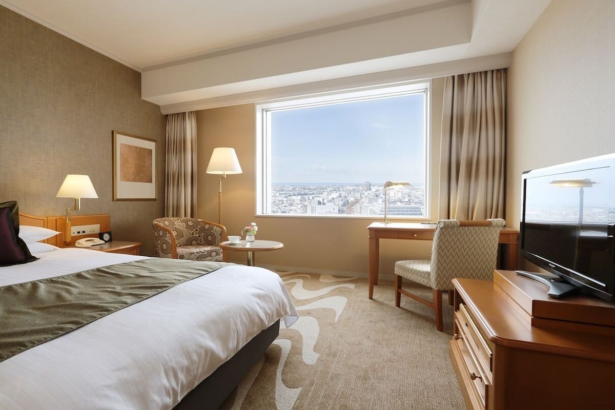 A hotel room with a bed, a desk, a chair, a TV, a window with a city view, and a lamp.
