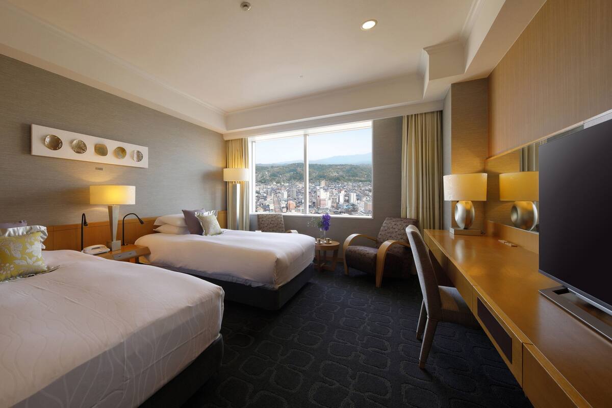 A hotel room with two beds, a desk, and a large window offering a city view.