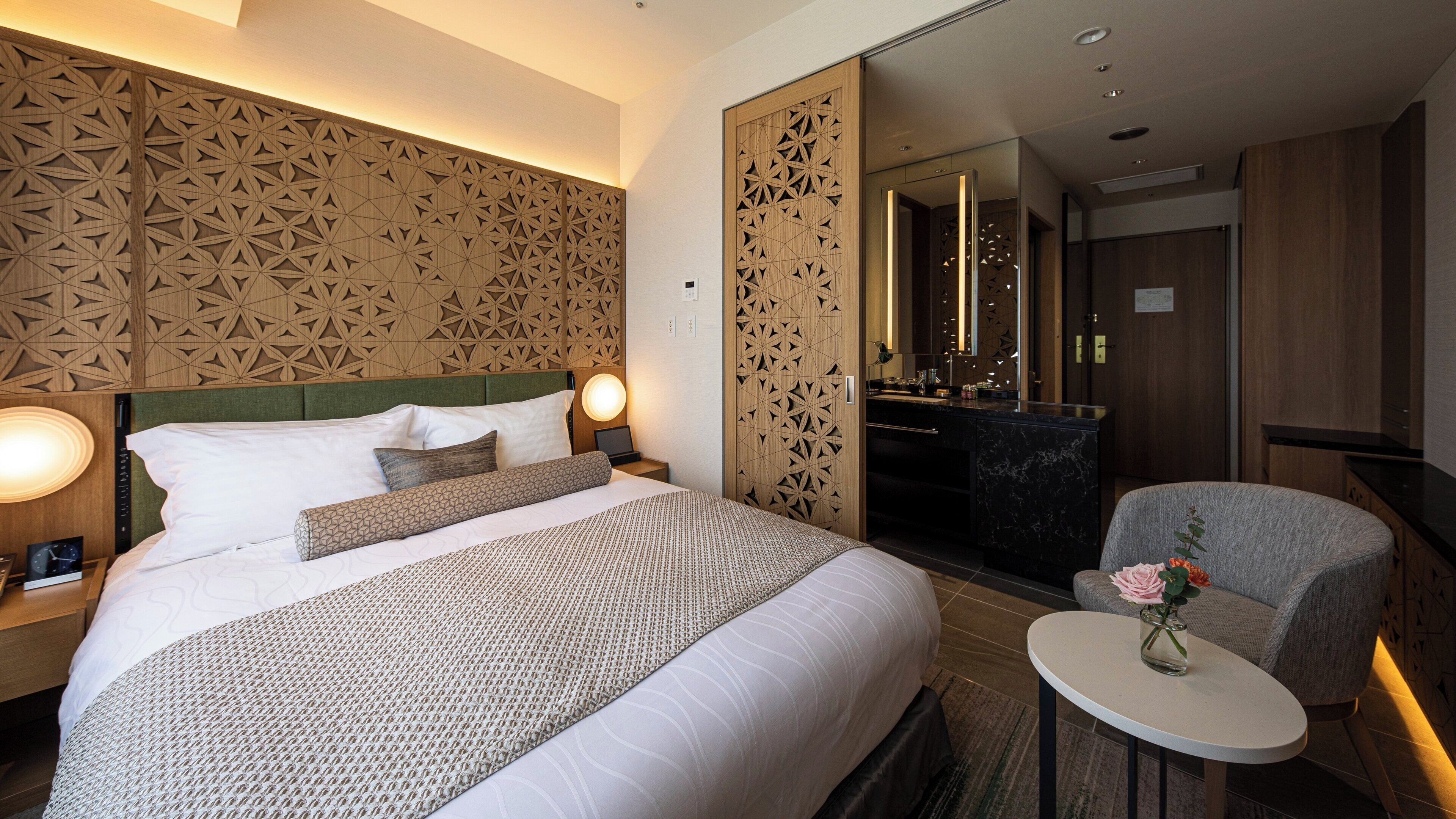 Habitación de hotel con una cama grande, un escritorio, una silla y un baño visible a través de una puerta corrediza.