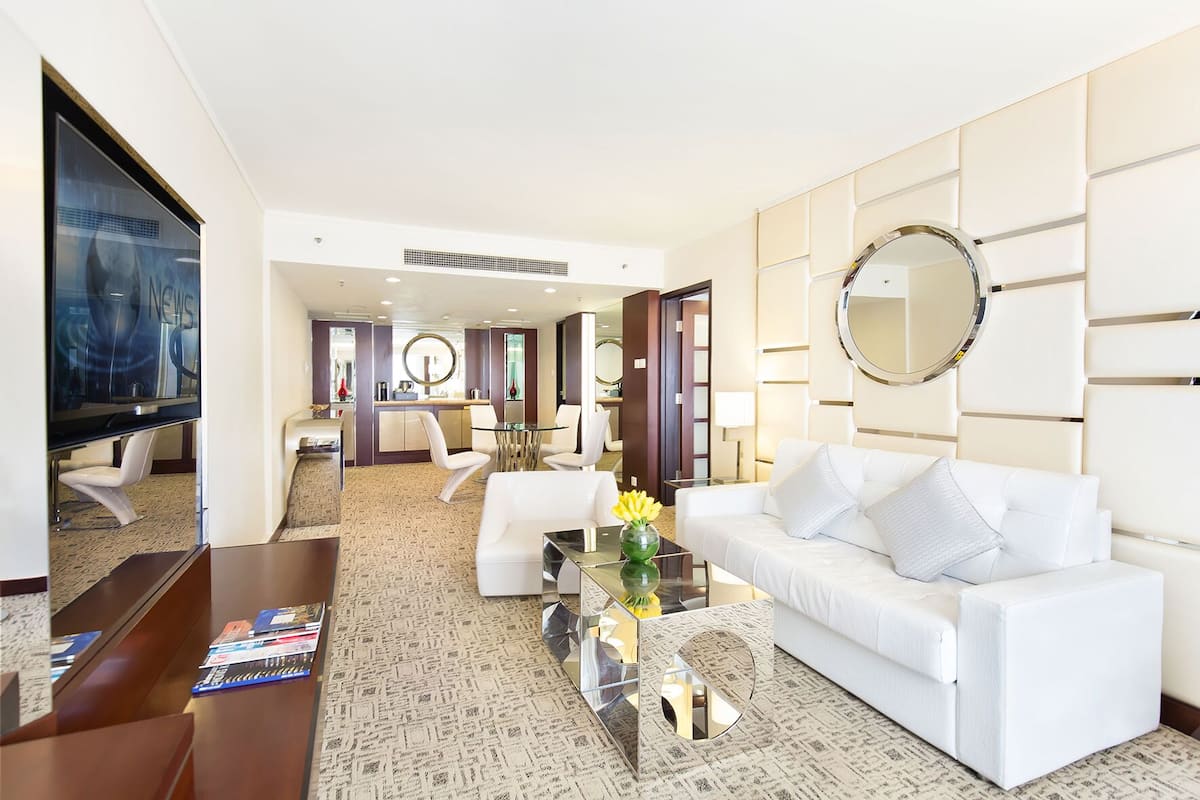 Una habitación de hotel moderna con un sofá blanco, una mesita de centro, un televisor y un área de comedor al fondo.